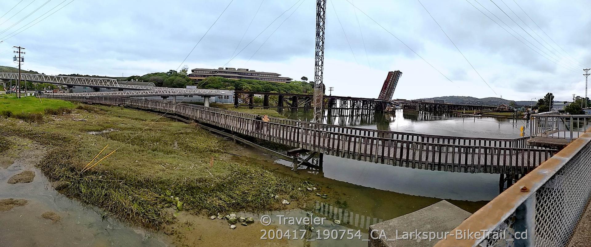 A long wooden boardwalk over muddy water, with bridges and buildings in distance.