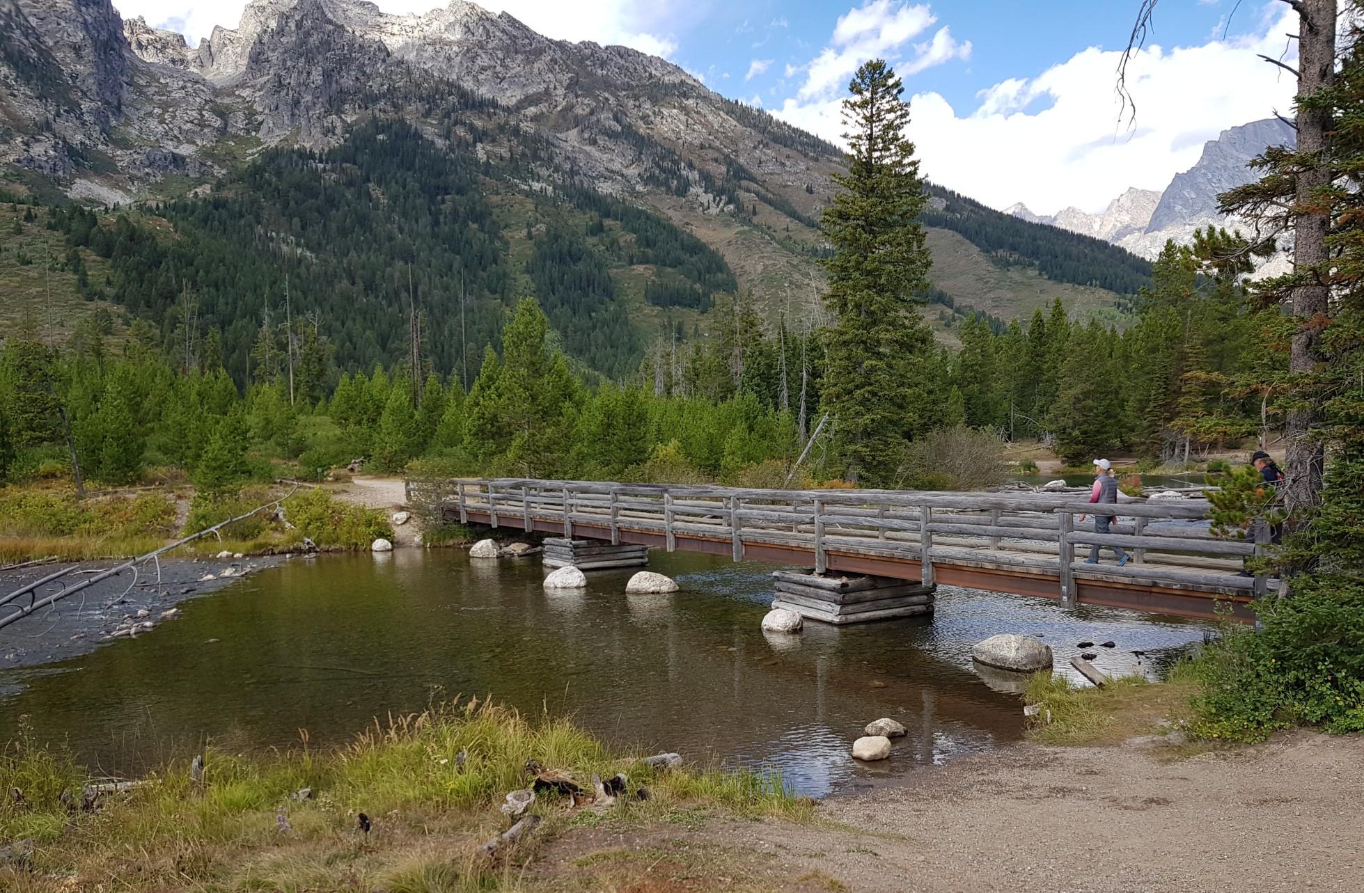 Two people cross a wooden bridge over a stream, surrounded by forest and mountains.