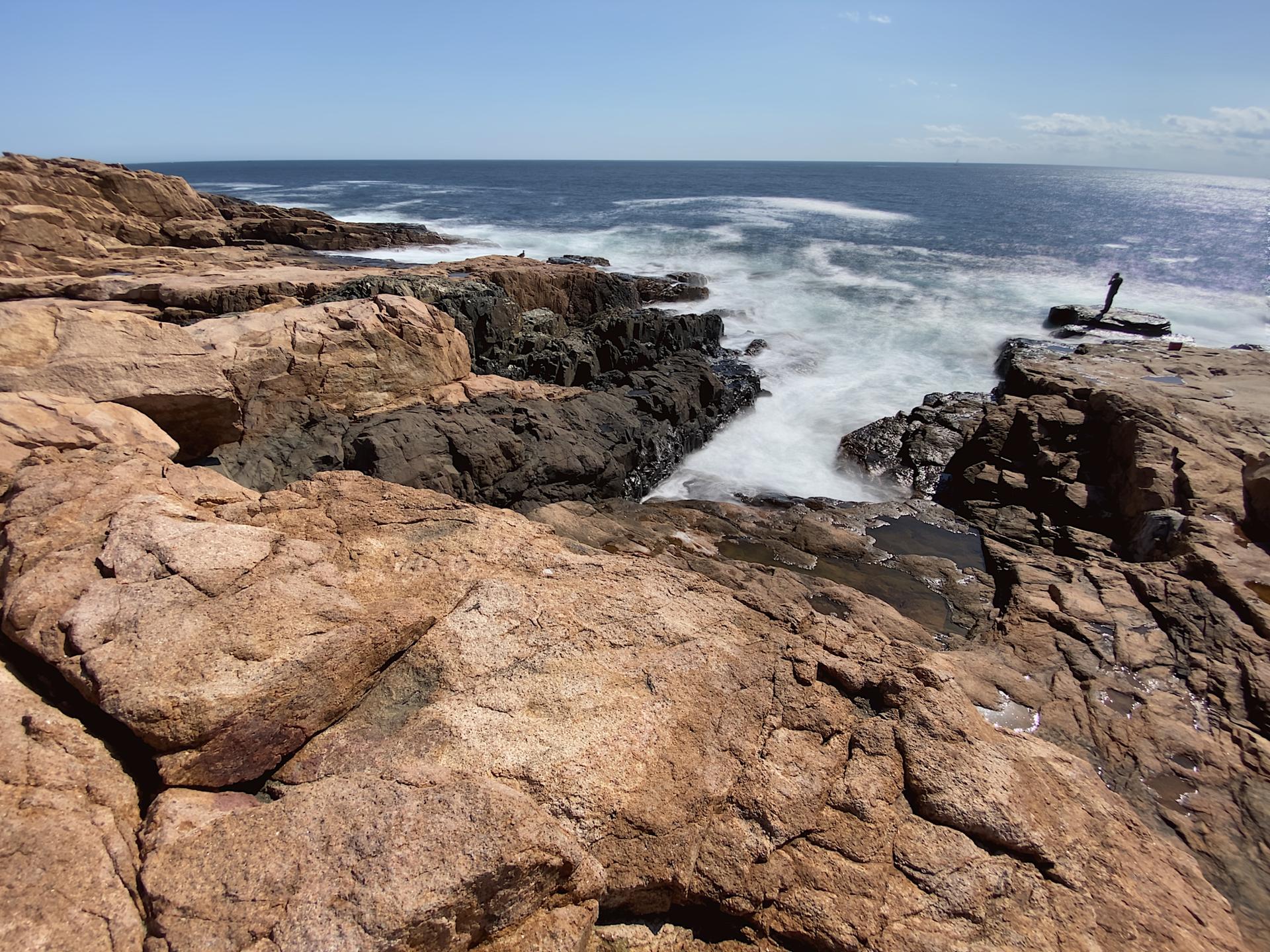 Rugged brown rocky coastline with white ocean waves crashing under a blue sky.