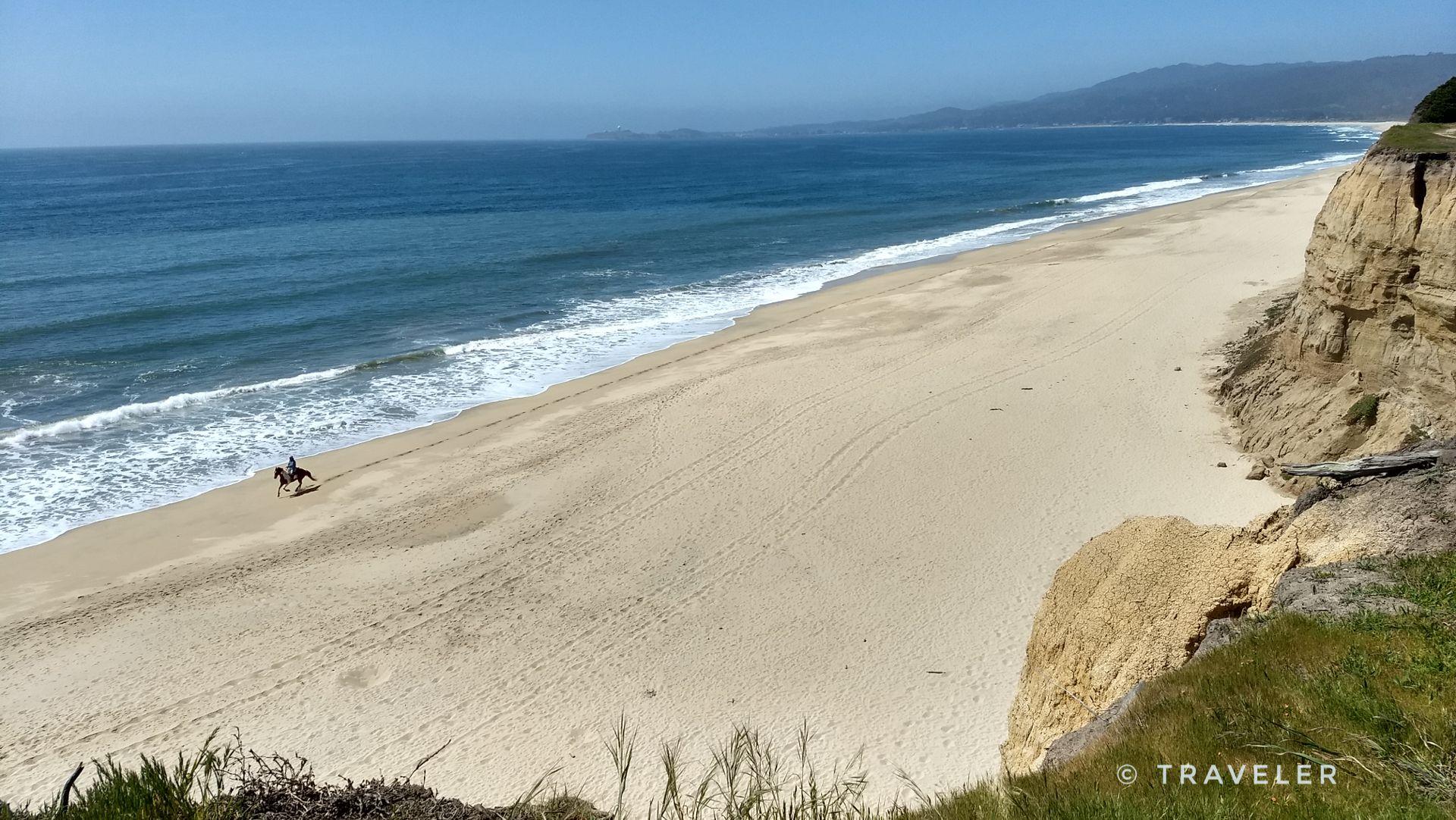 Sunny wide beach with ocean waves, a person riding a horse.
