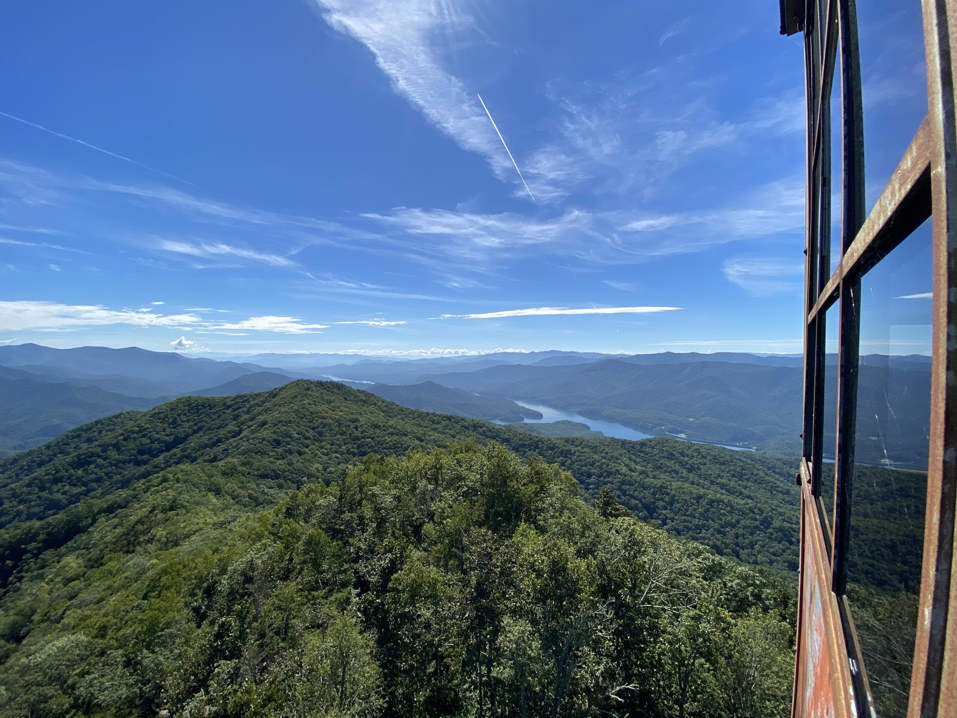Panoramic view of green forested mountains and a distant lake from a high vantage point.