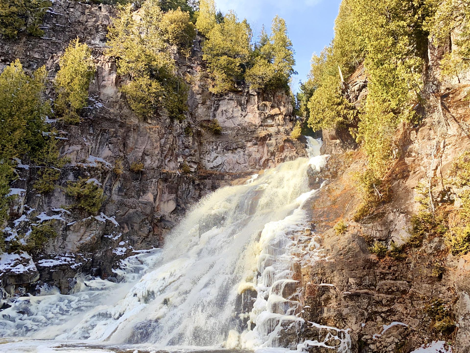 Powerful waterfall flows over icy rocks in a sunlit, forested gorge.