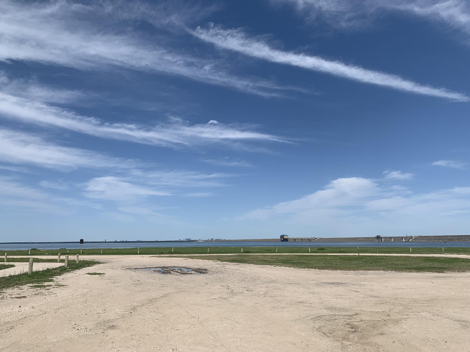 Dirt foreground, grassy bank, and calm water under a bright, cloudy sky.