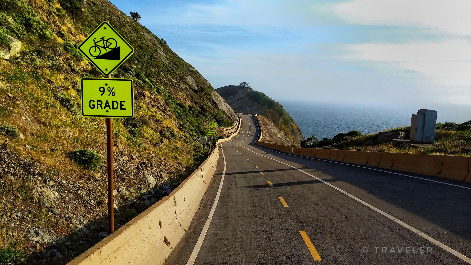 Winding coastal road with steep grade signs, ocean, and sunny sky.