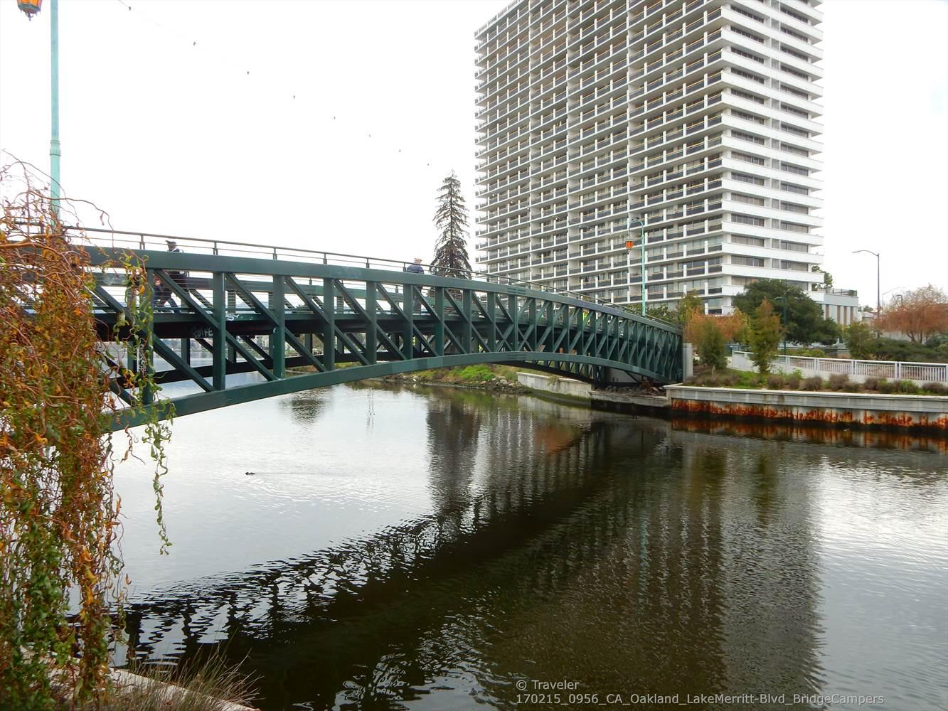 A green truss bridge over water, reflecting a tall white building.