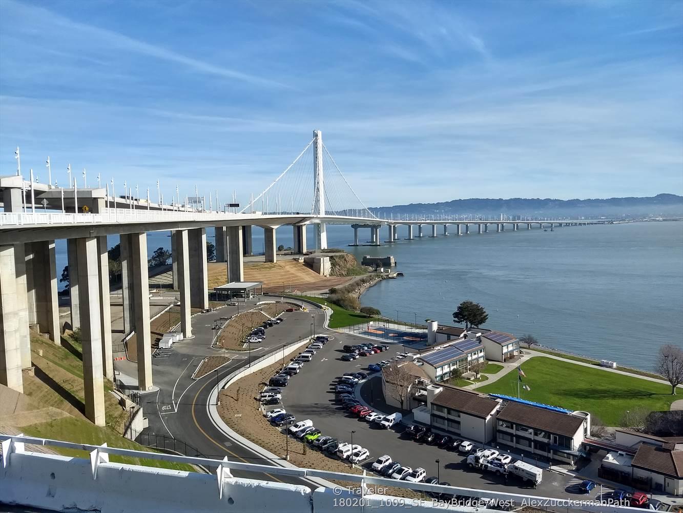 Large suspension bridge over water, with a parking lot and buildings below. Bright day.