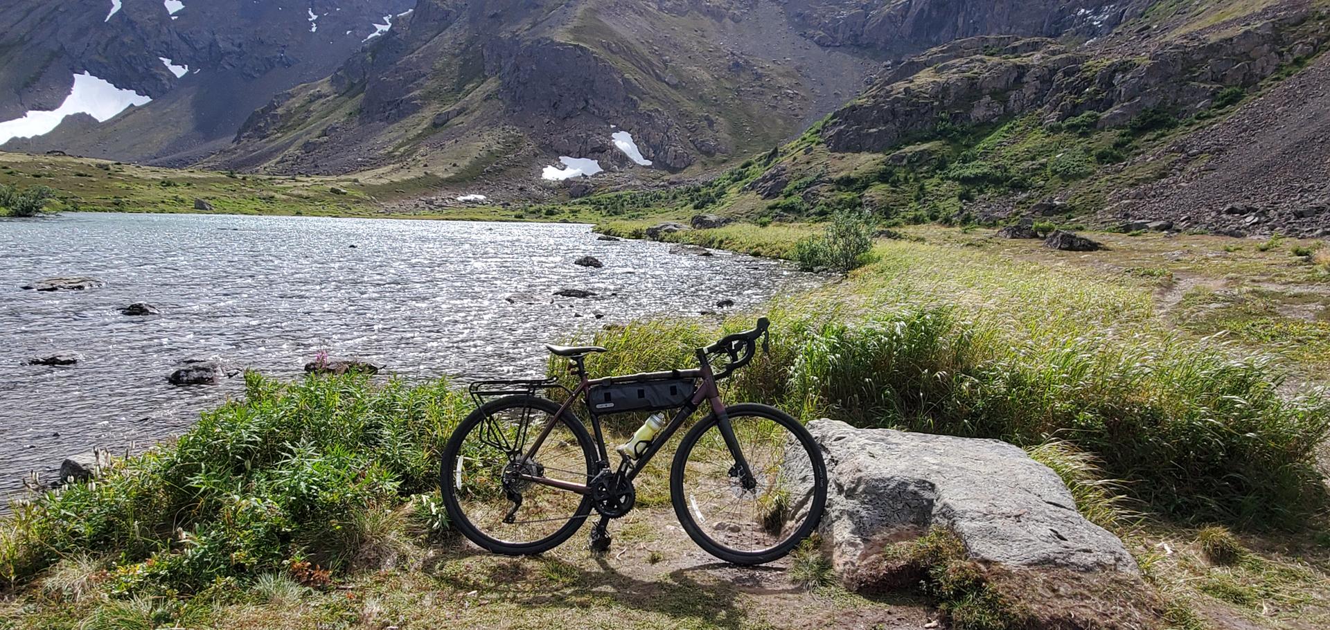 A touring bicycle rests by a mountain lake under bright daylight.