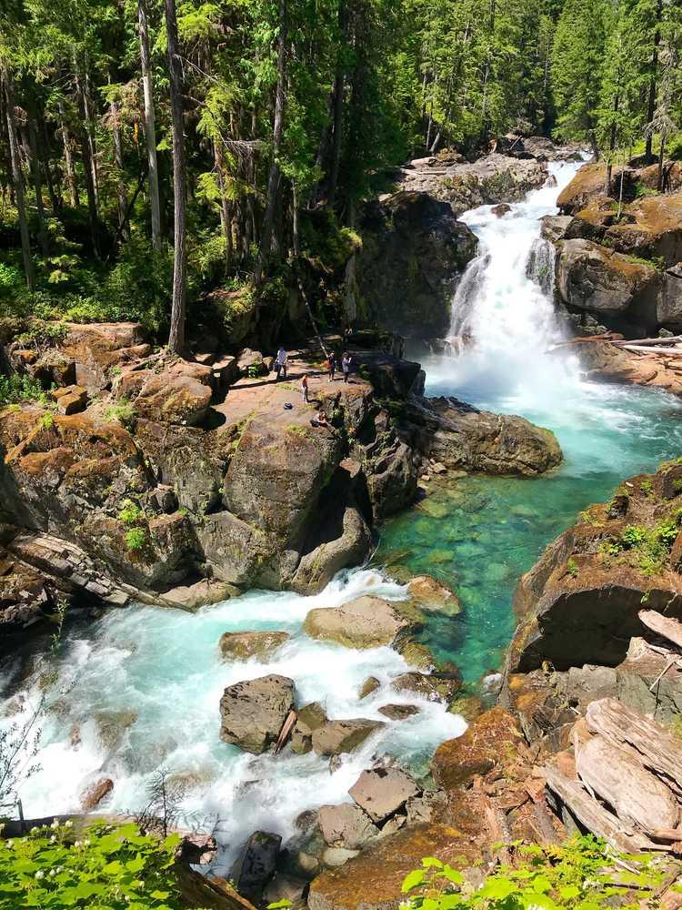 People on rocks by a powerful waterfall and clear river in a sunny evergreen forest.