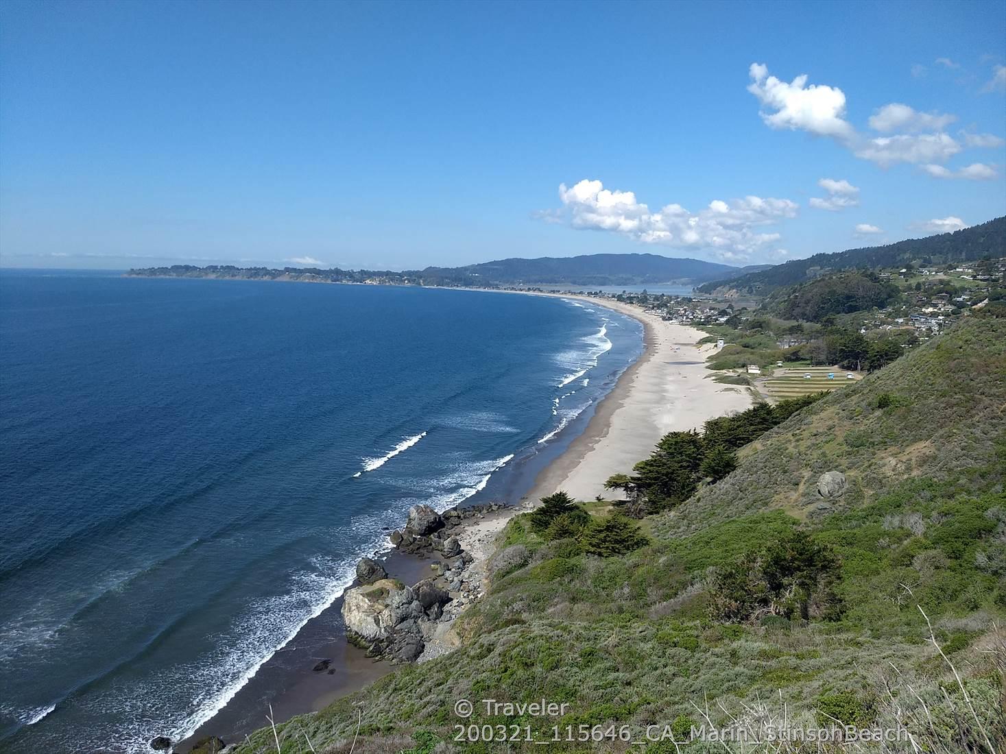 Panoramic view of a long sandy beach, ocean waves, and green coastal cliffs.