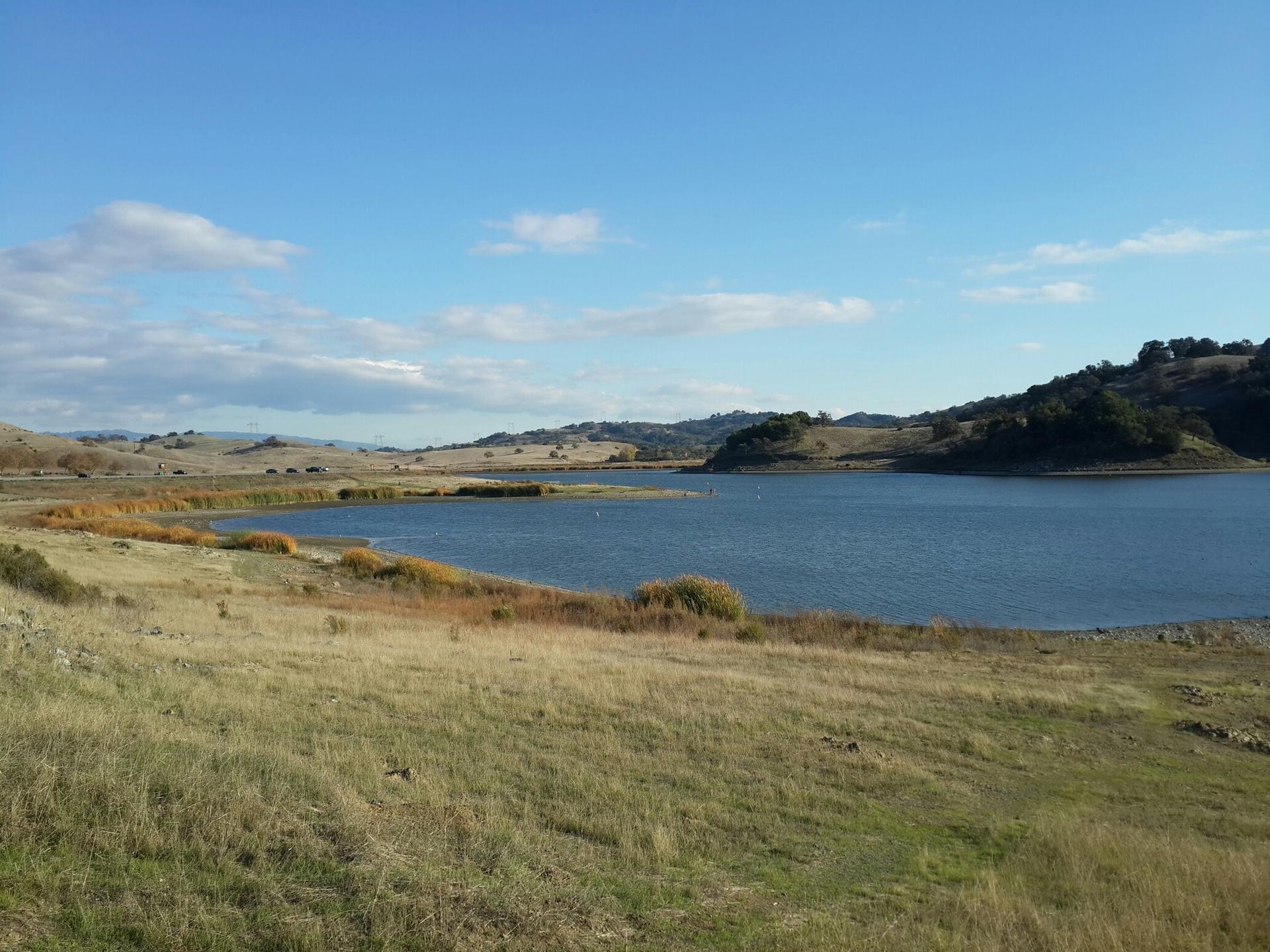 Wide lake surrounded by dry and green hills under a partly cloudy sky.
