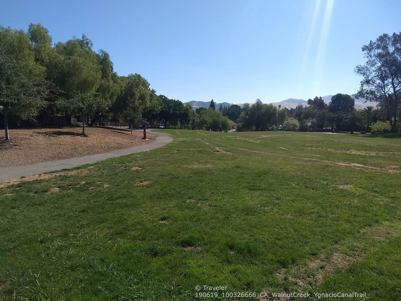 A wide, sunny green field with a paved path curving left, distant hills.