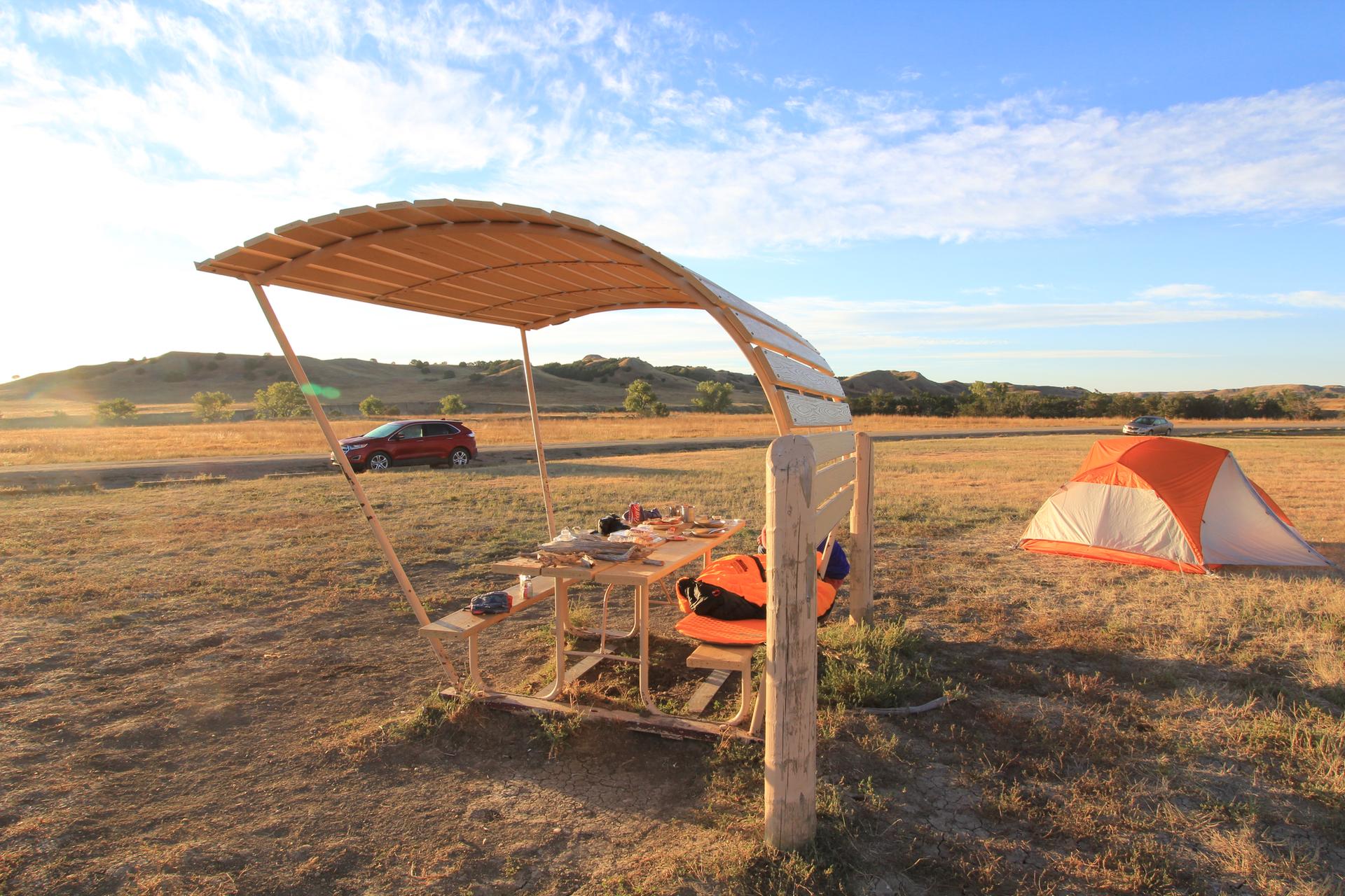 Camping setup with wooden shelter, orange tent, and red car in sunny field.