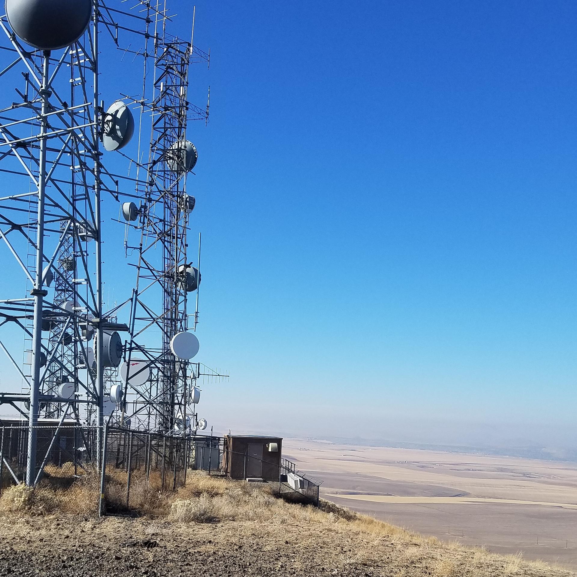 Communication towers on a dry hill overlooking a vast plain under a clear blue sky.