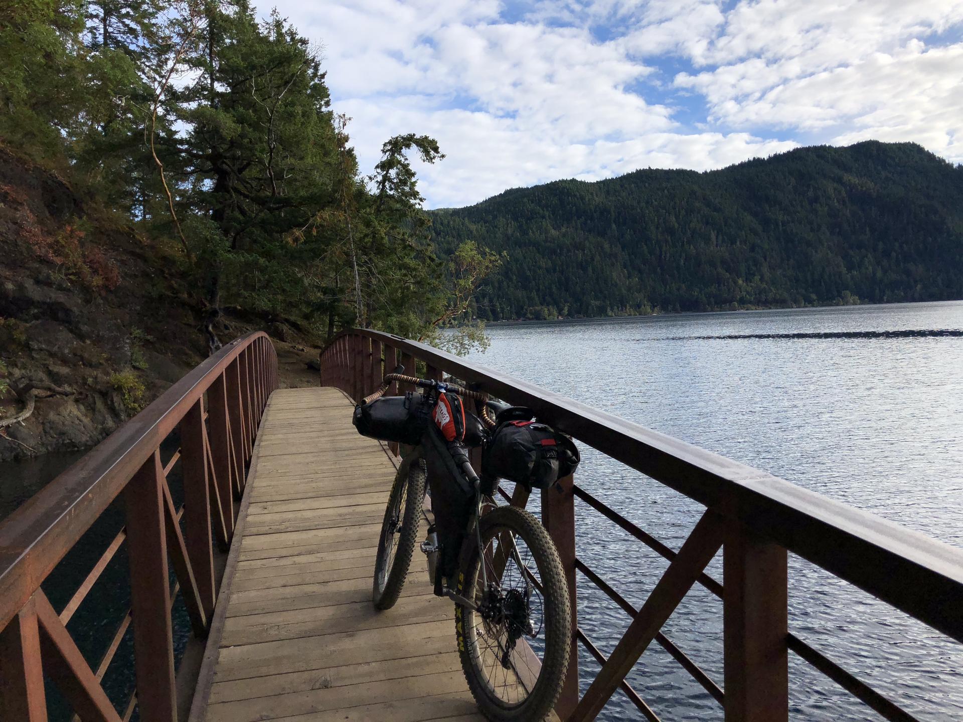 Bicycle parked on a wooden bridge overlooking a calm lake and forested hills.