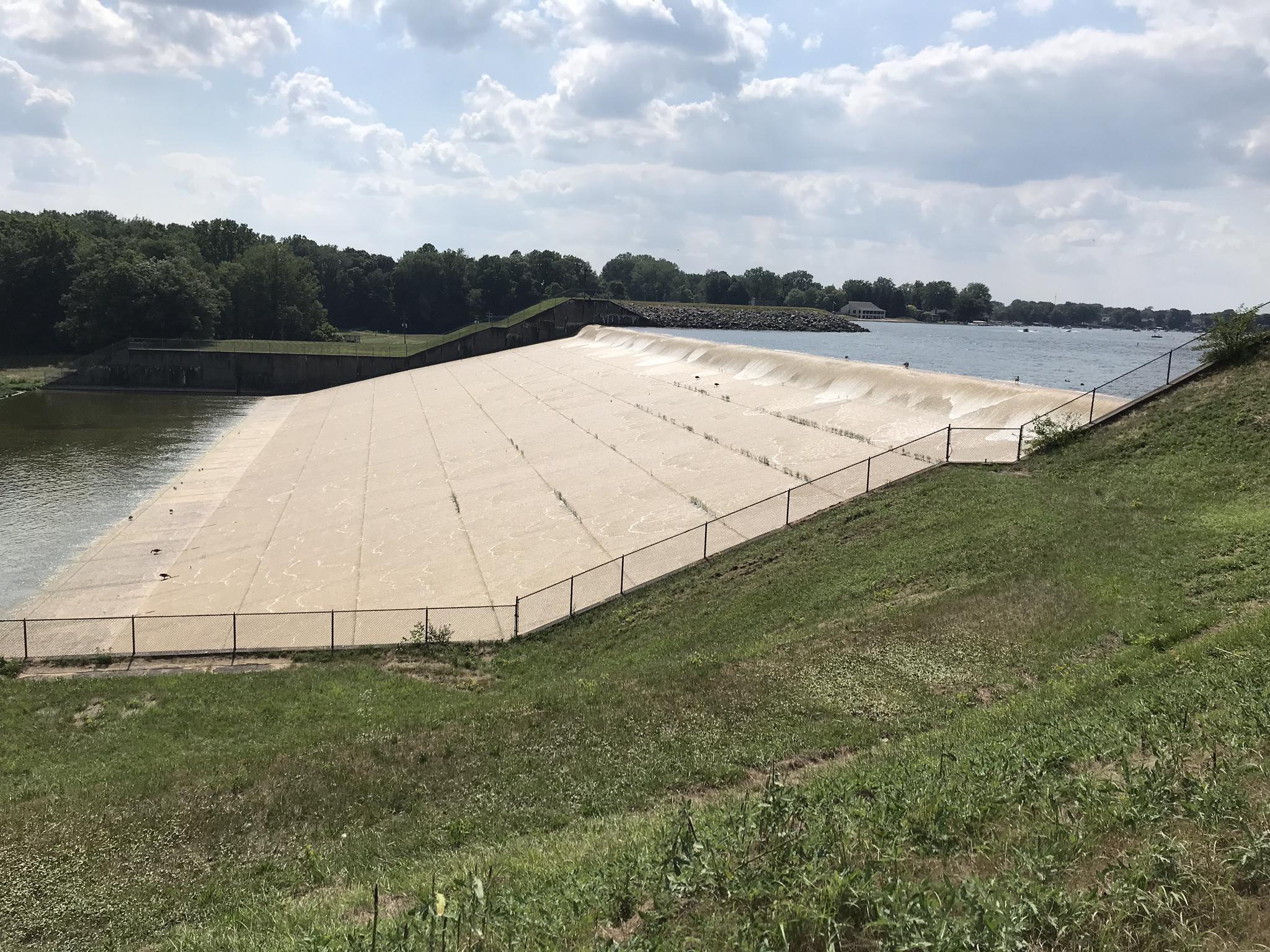 Wide concrete spillway with water flowing, surrounded by green banks.