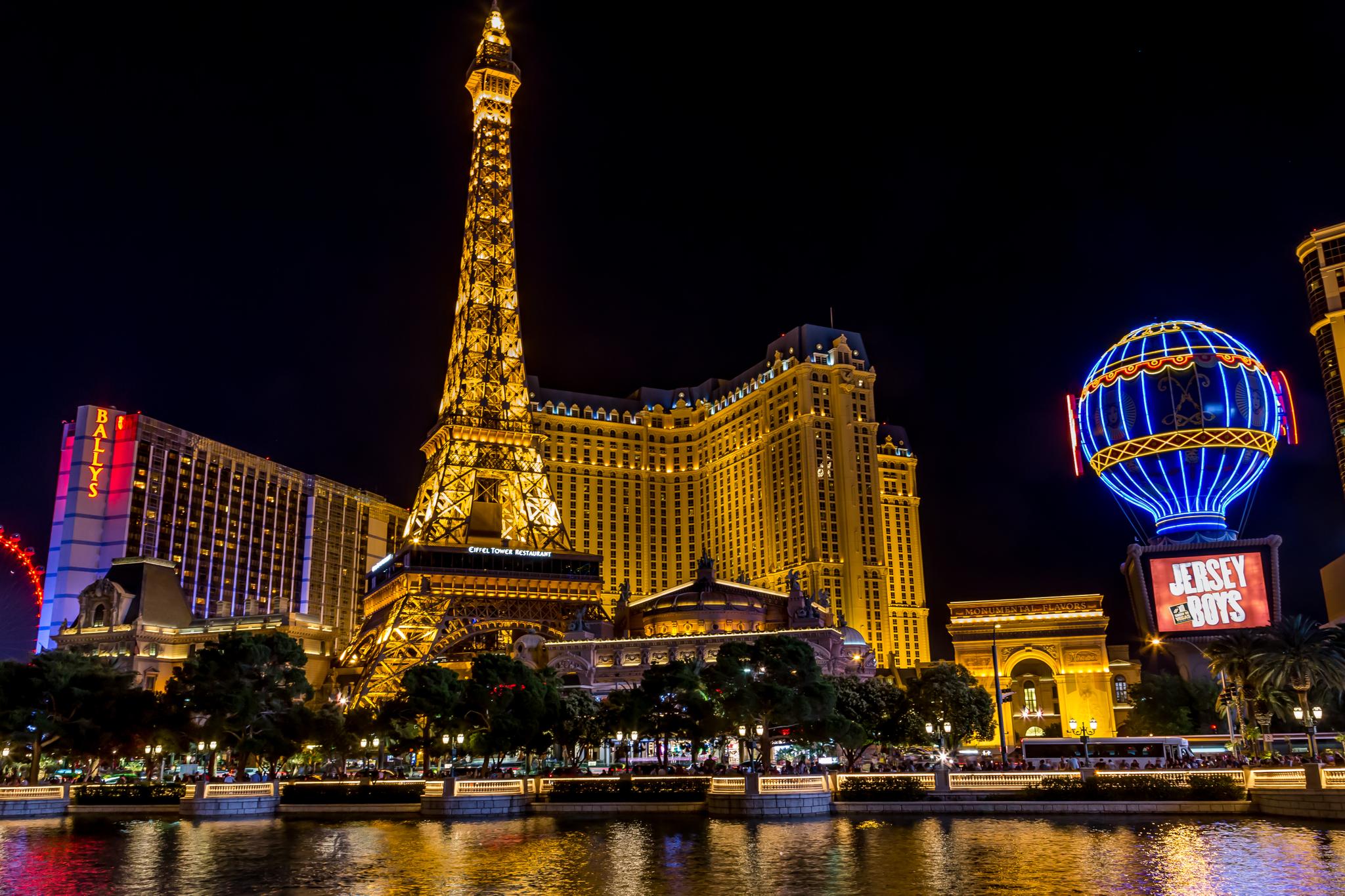 Brightly lit replica tower and buildings at night, reflecting in water.
