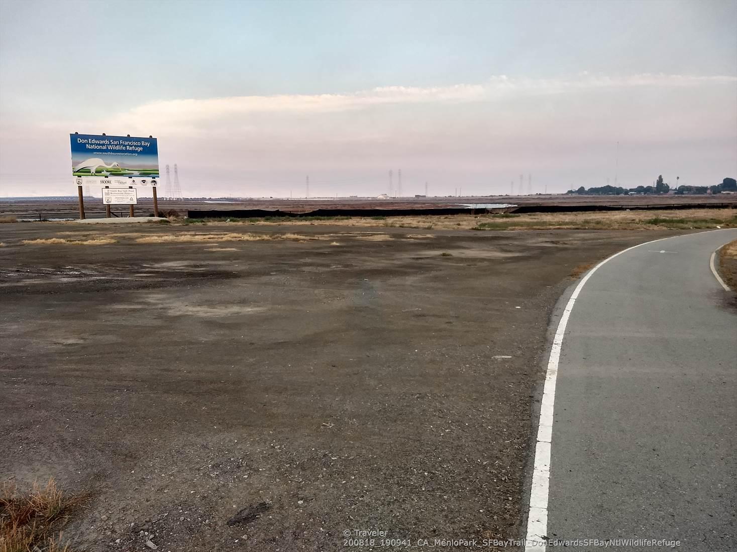 A paved path curves right next to a large dirt area and distant sign.