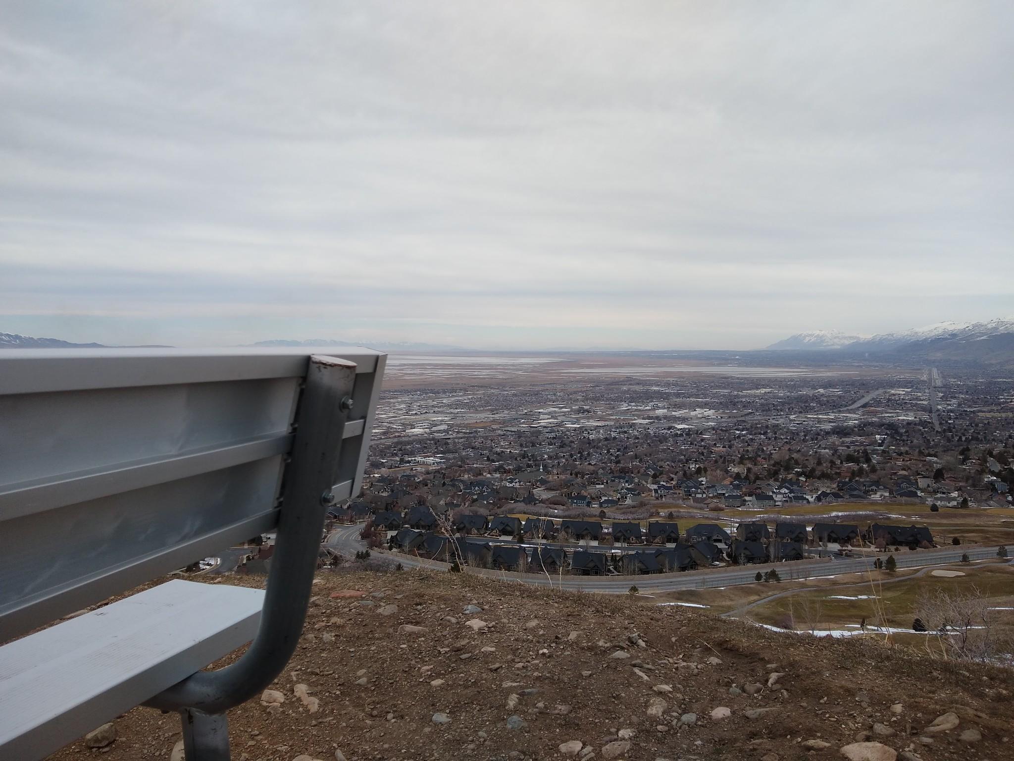 Metal bench overlooks a sprawling town and distant mountains under an overcast sky.