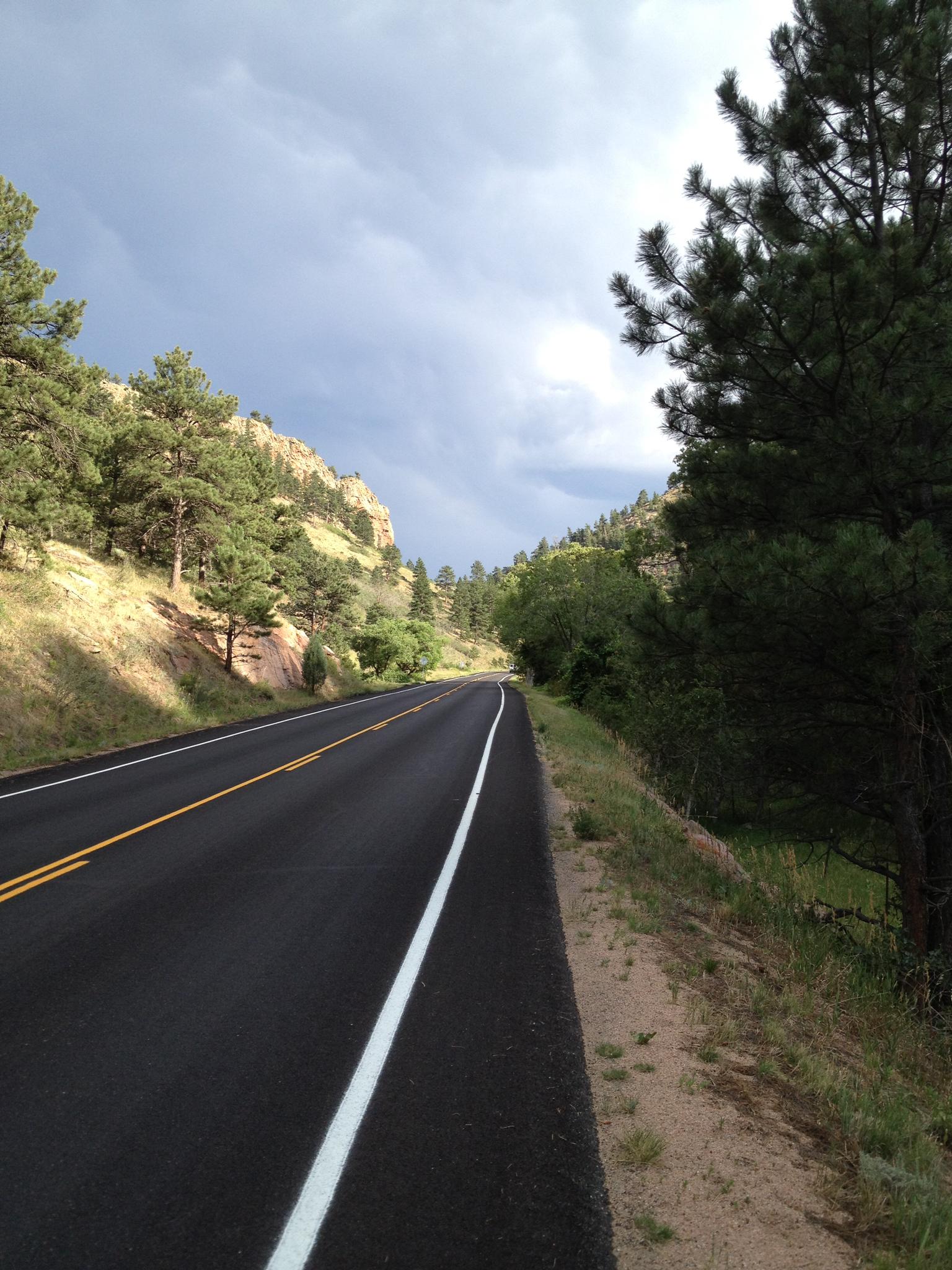Paved road winding through a forested mountain pass under dramatic clouds.