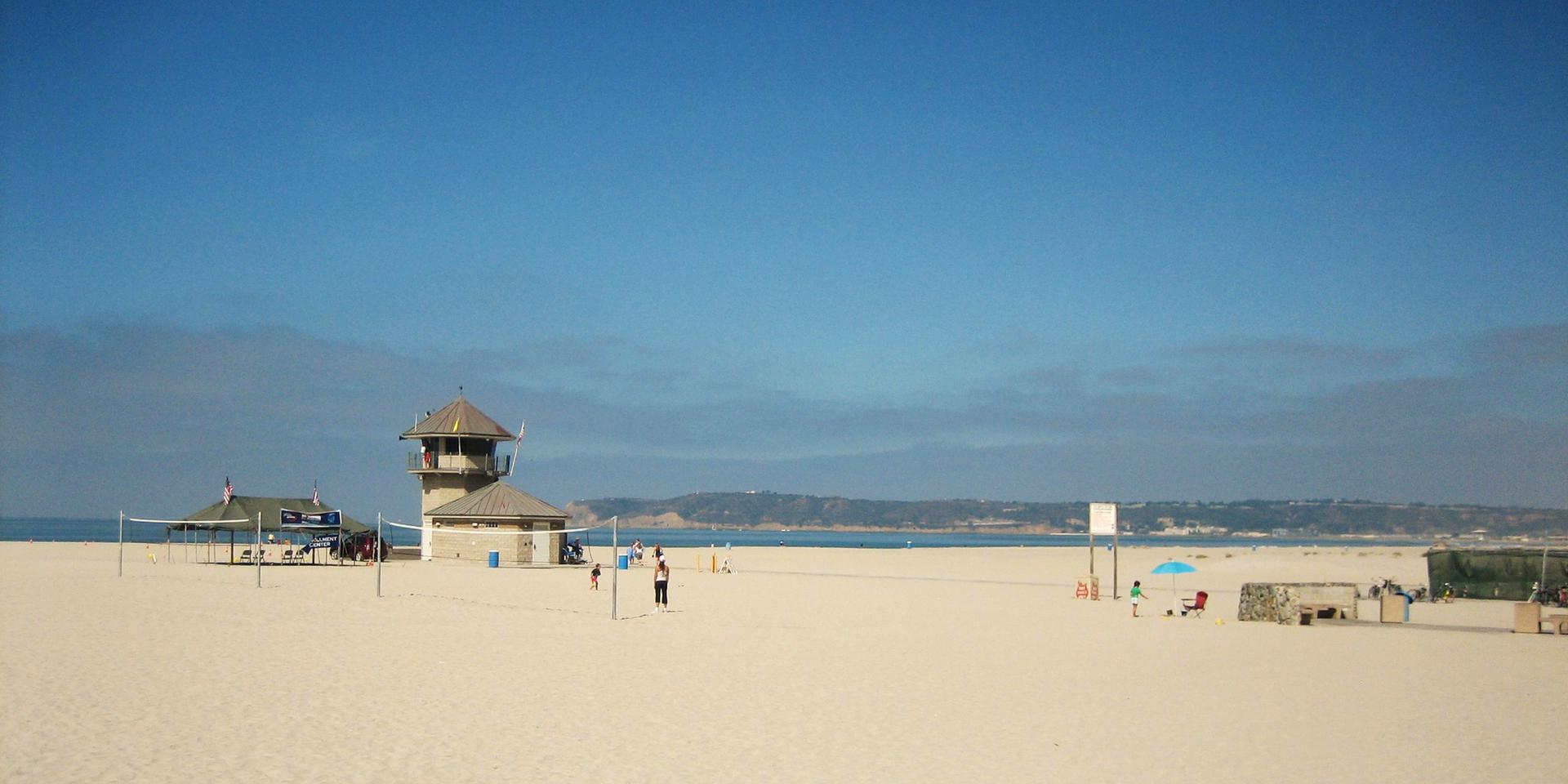 Sandy beach with a lifeguard tower, volleyball net, and people playing under clear sky.