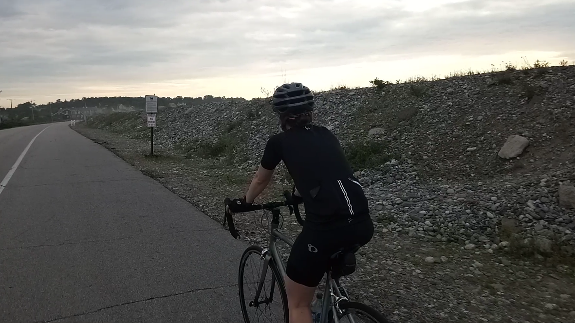 Person cycling on a paved road next to a gravel embankment under an overcast sky.