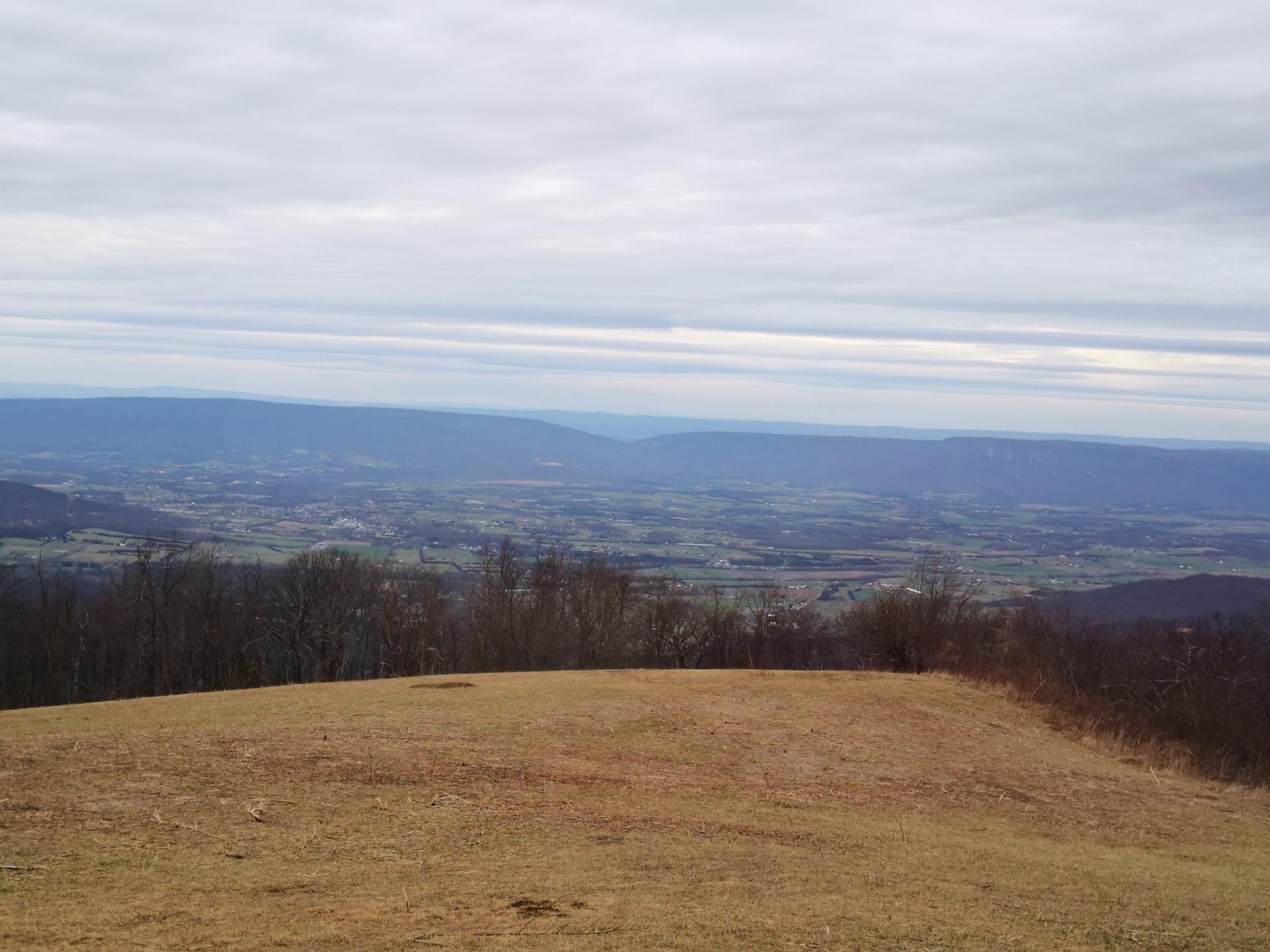 Overcast view of a wide valley from a grassy, brown hilltop.