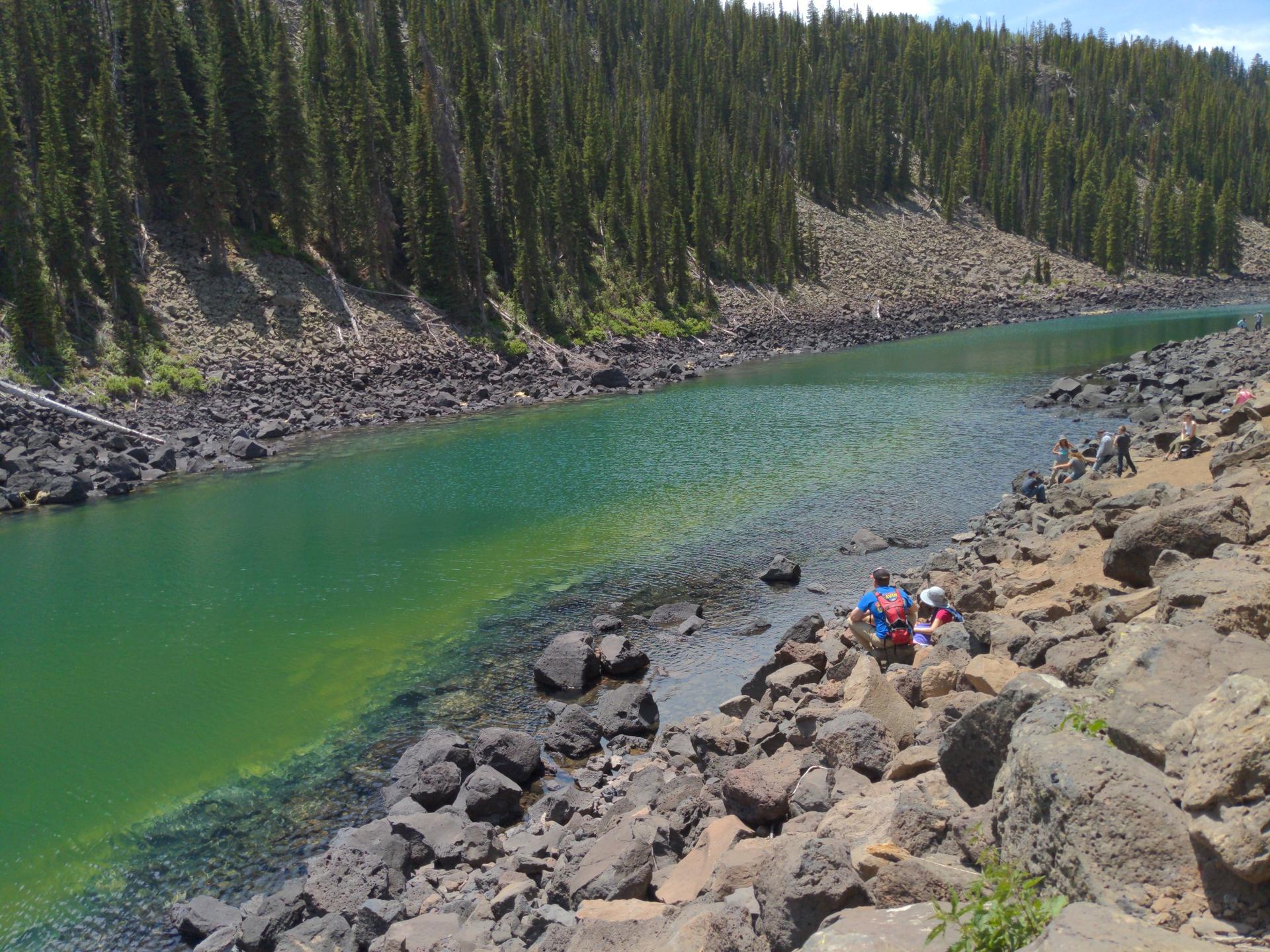People sitting on rocky shore next to a green lake with forested hills.