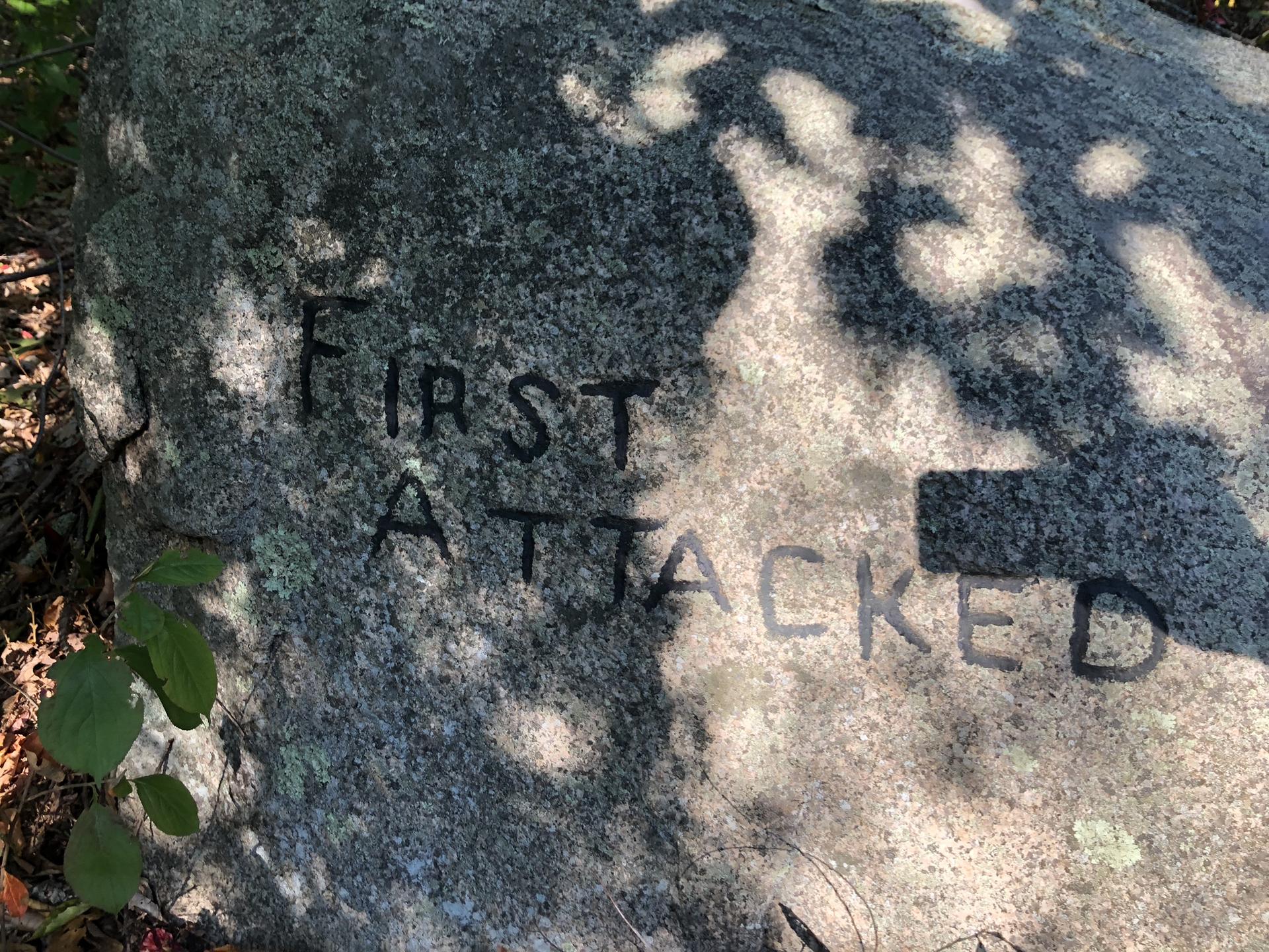 Close-up of a gray rock with "FIRST ATTACKED" carved, dappled sunlight.