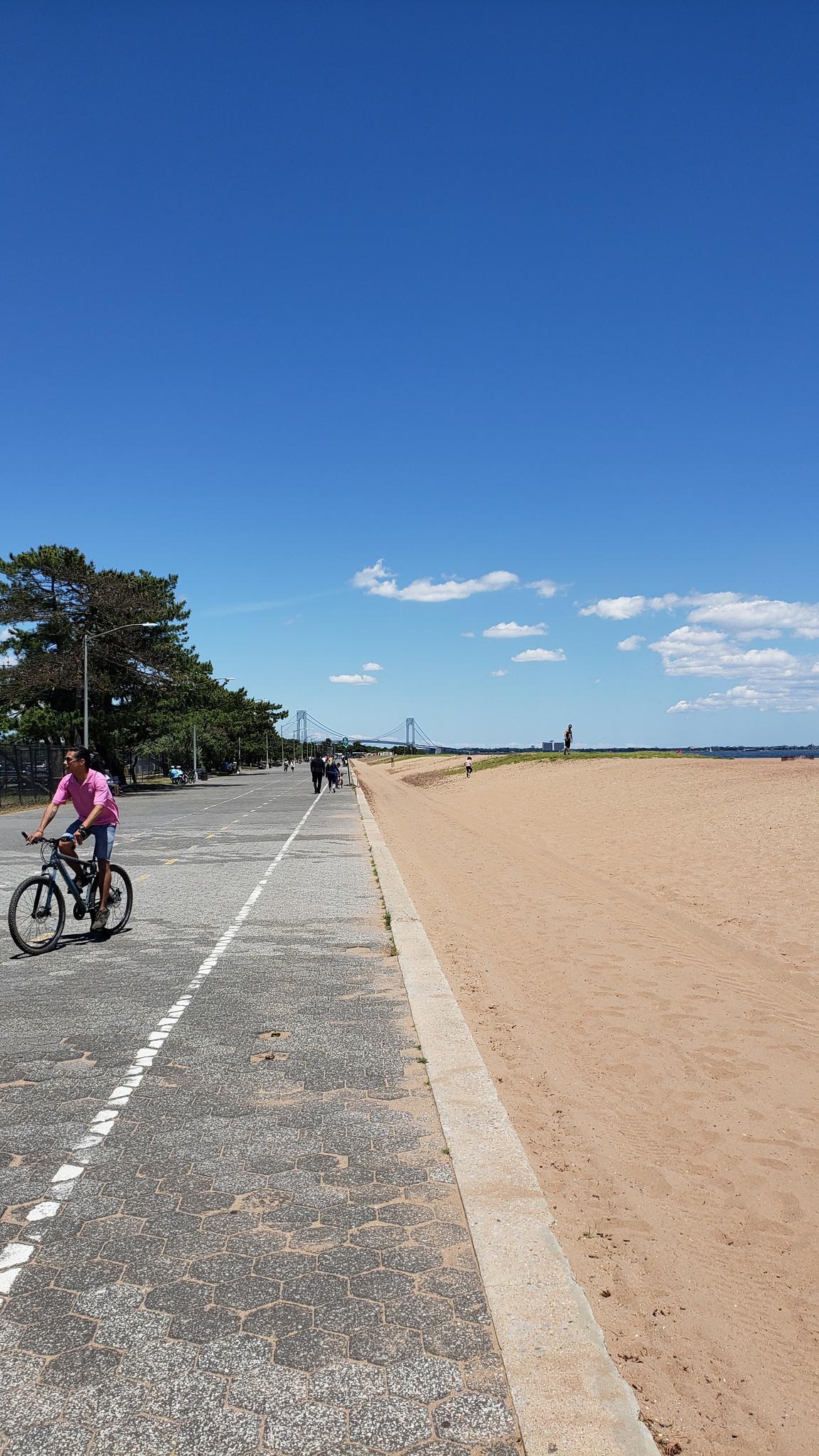 Person cycling on a paved path next to a sandy beach under blue sky.