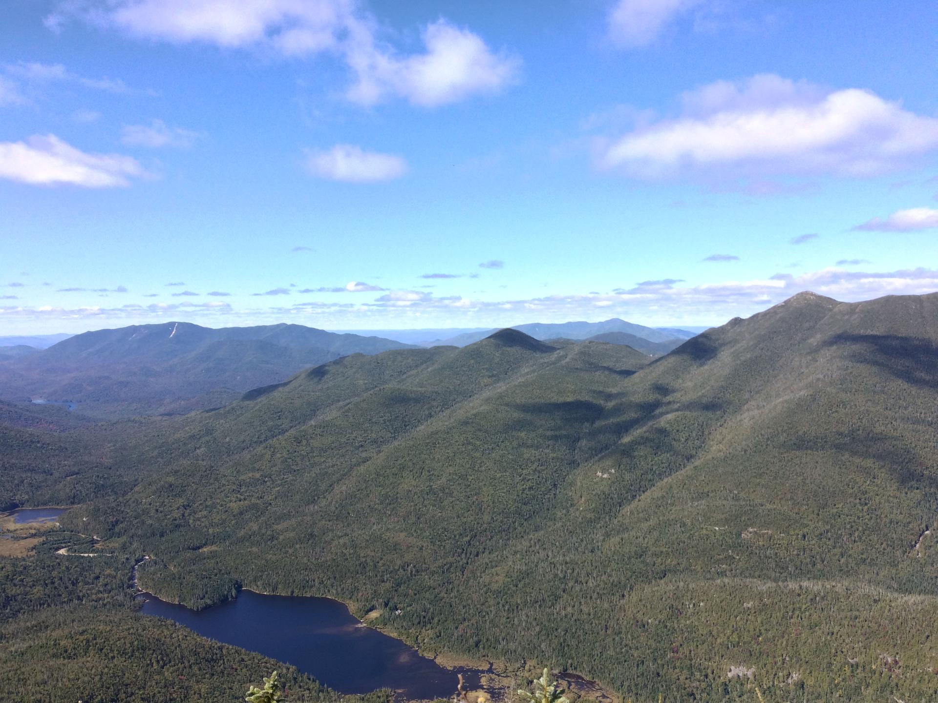 An expansive aerial view of forested mountains and a dark lake on a sunny day.