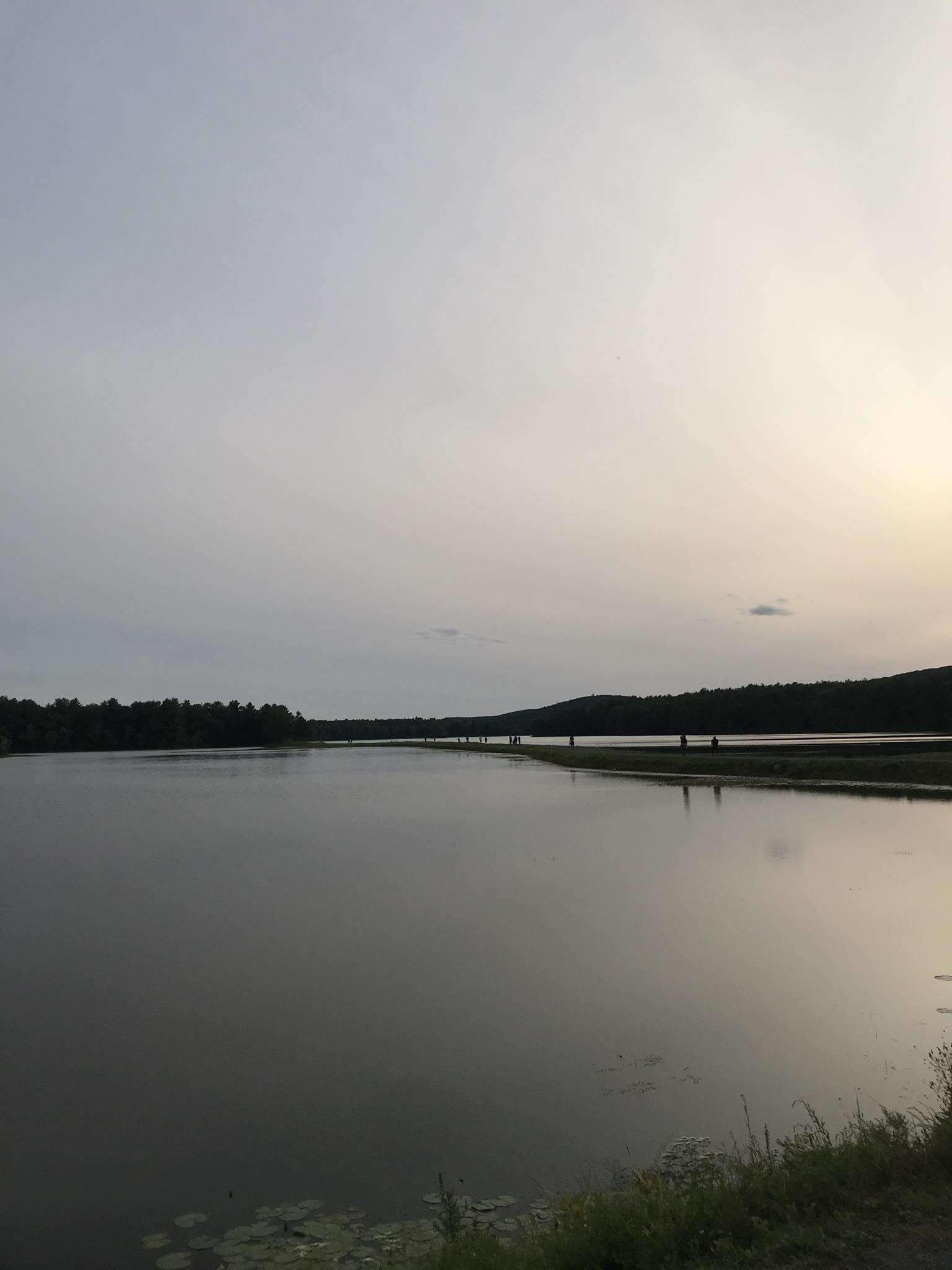 Wide, calm lake under a pale sky with distant forested hills and people.