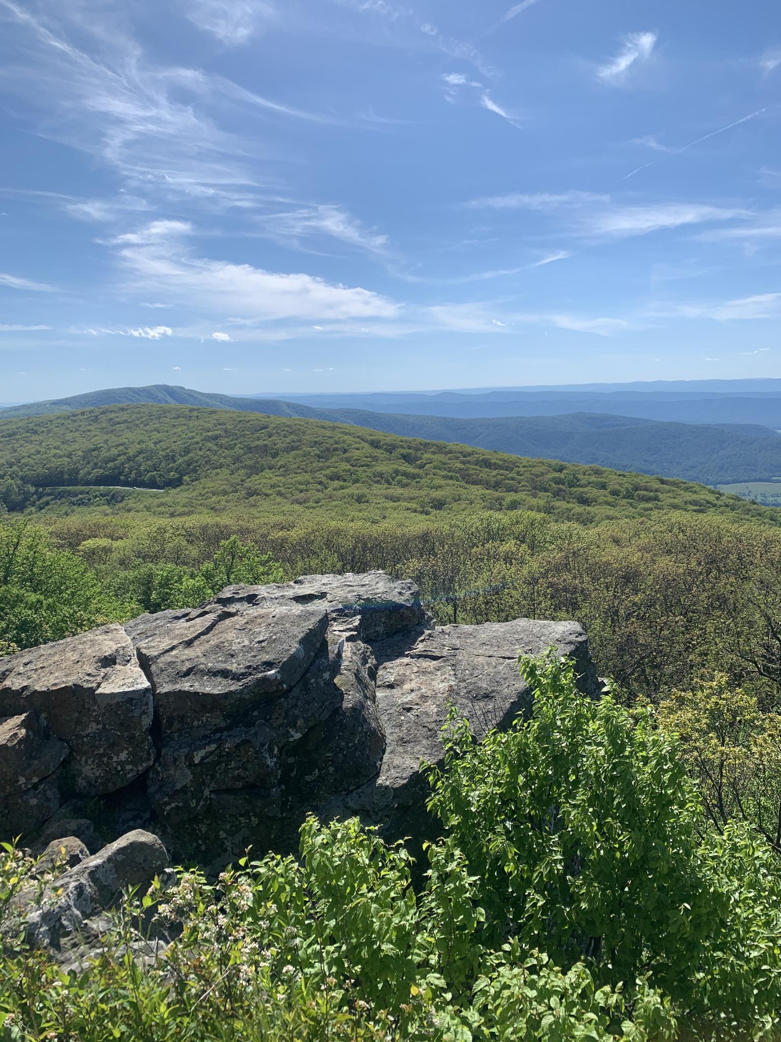 Rocky outcrop overlooking verdant mountain ridges under a bright blue sky.