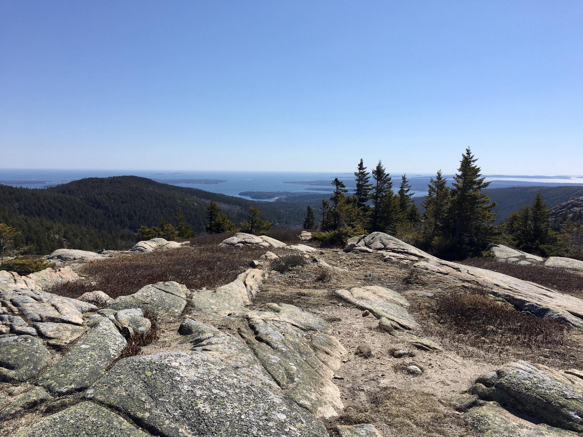 Rocky mountain summit with distant forested hills and ocean under clear sky.