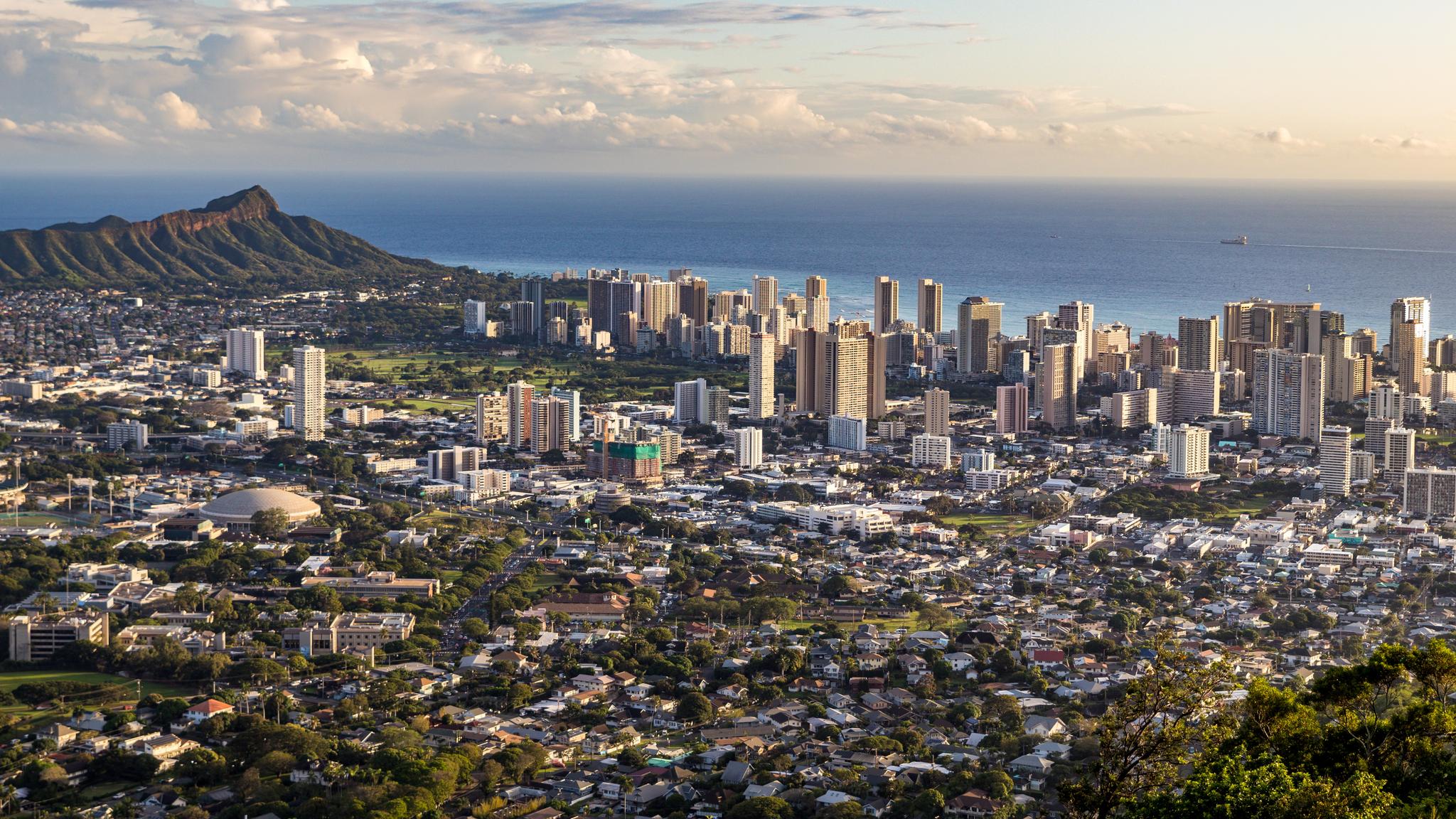 Panoramic view of a coastal city and mountain under warm, partly cloudy skies.