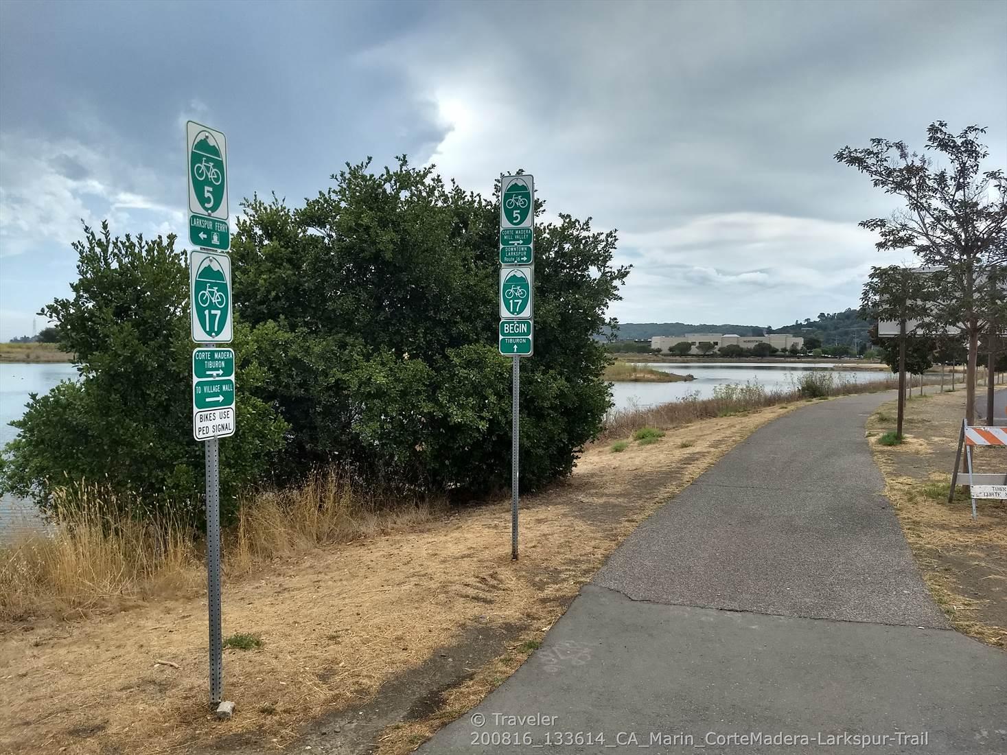 Two green trail signs stand beside a paved path and water.