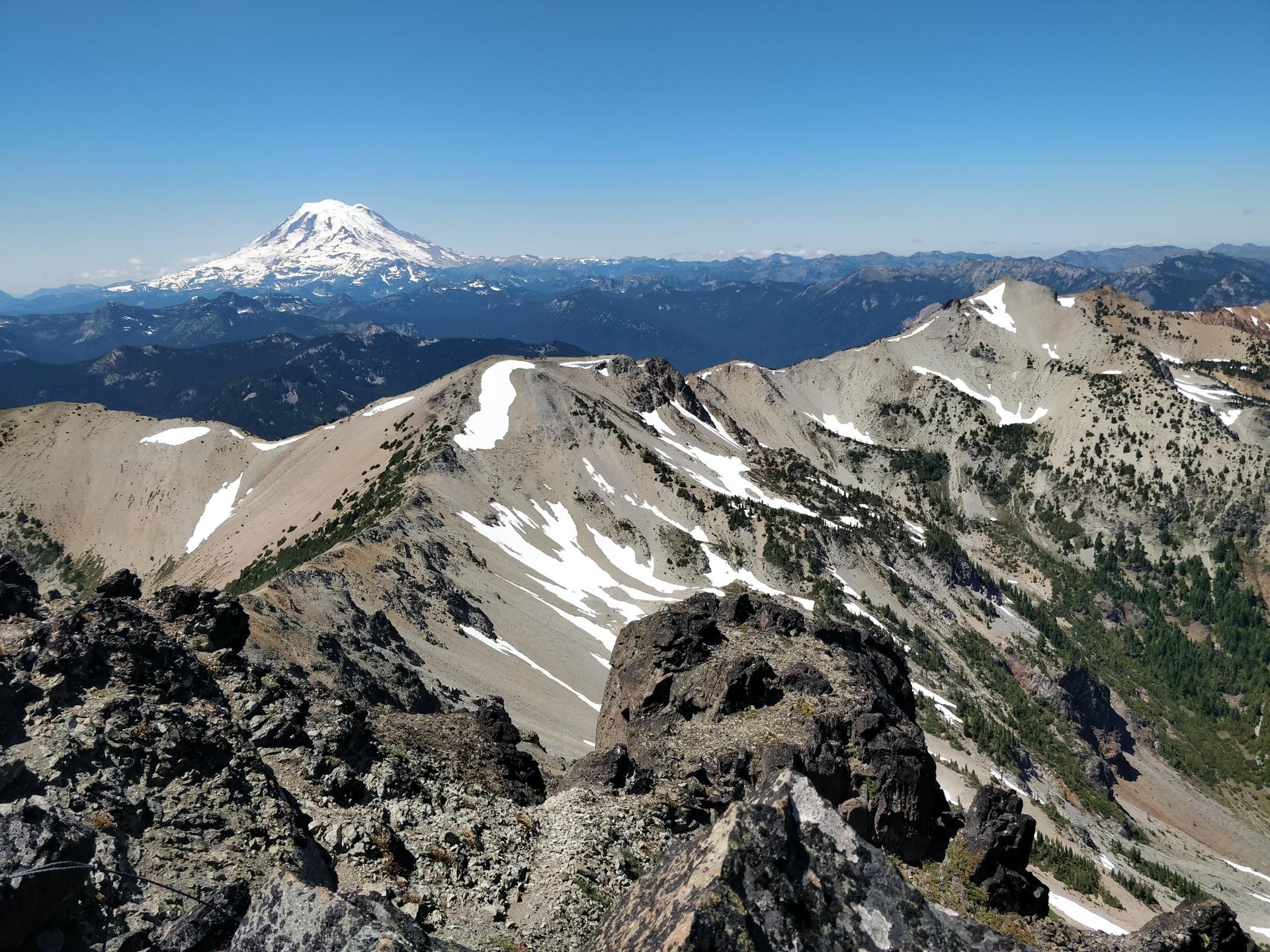 Rocky mountain peaks with snow patches, a distant snow-capped volcano, under bright blue sky.