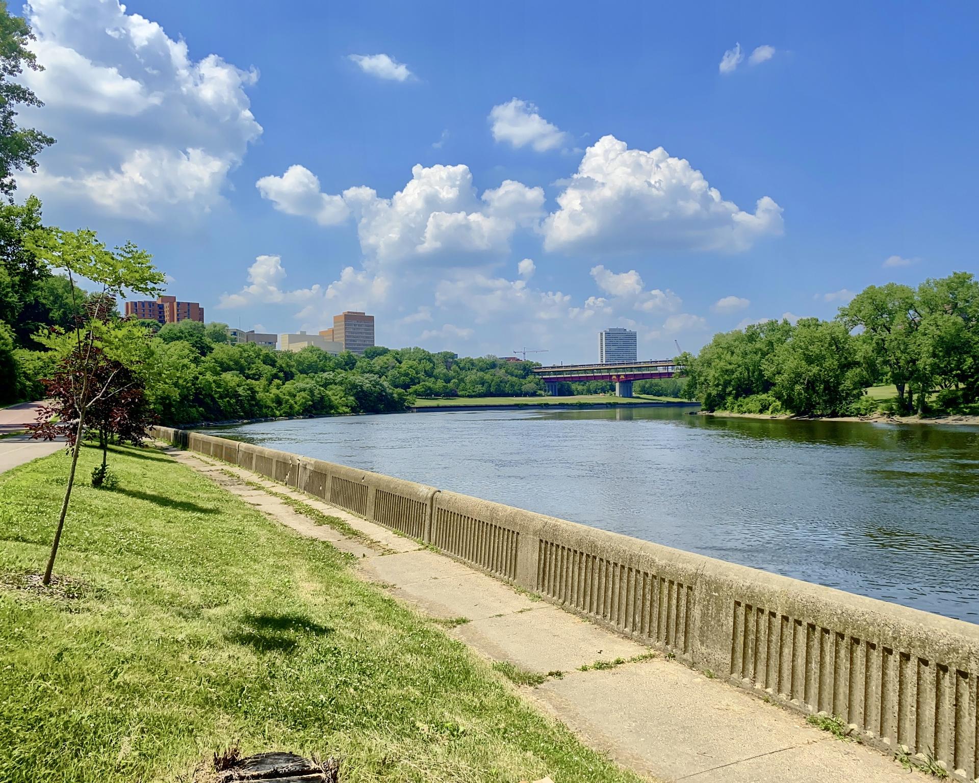 River flowing past a concrete path, green grass, and distant city buildings.