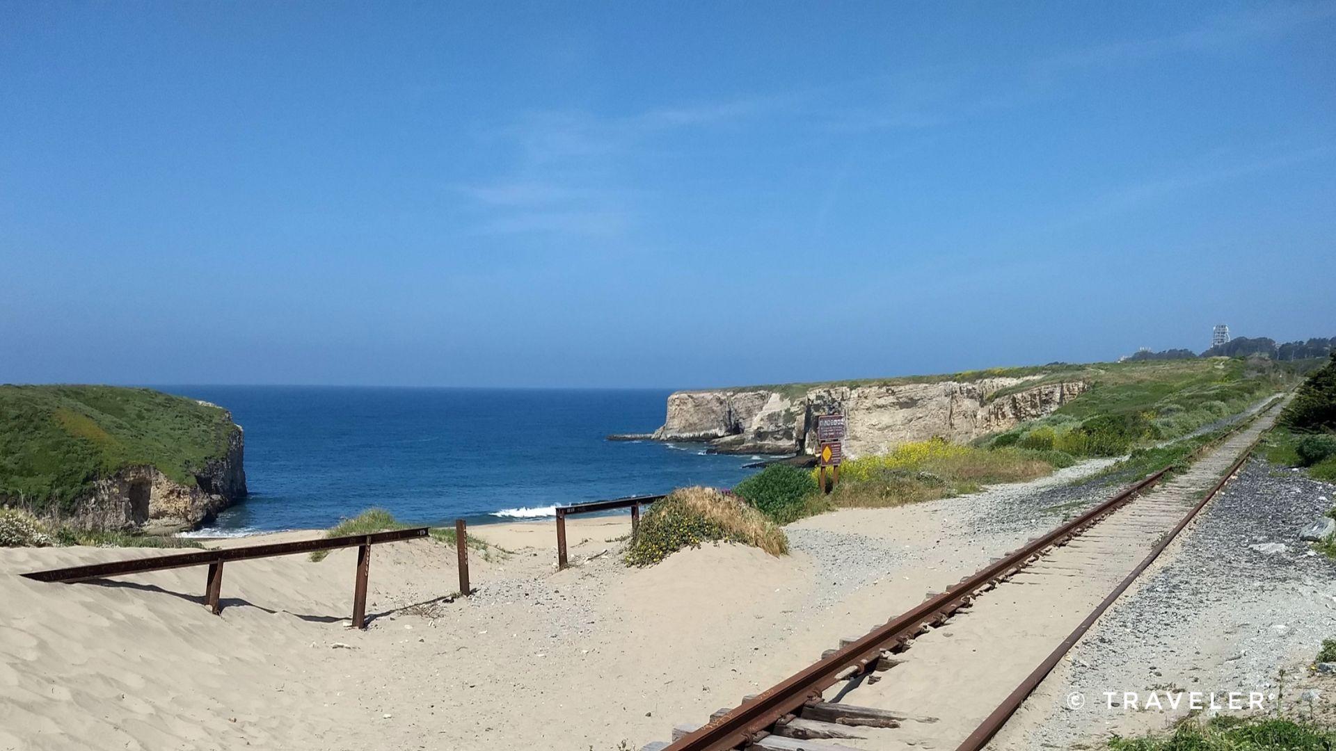 Sunny coastal scene with train tracks on sand, ocean, and distant cliffs.