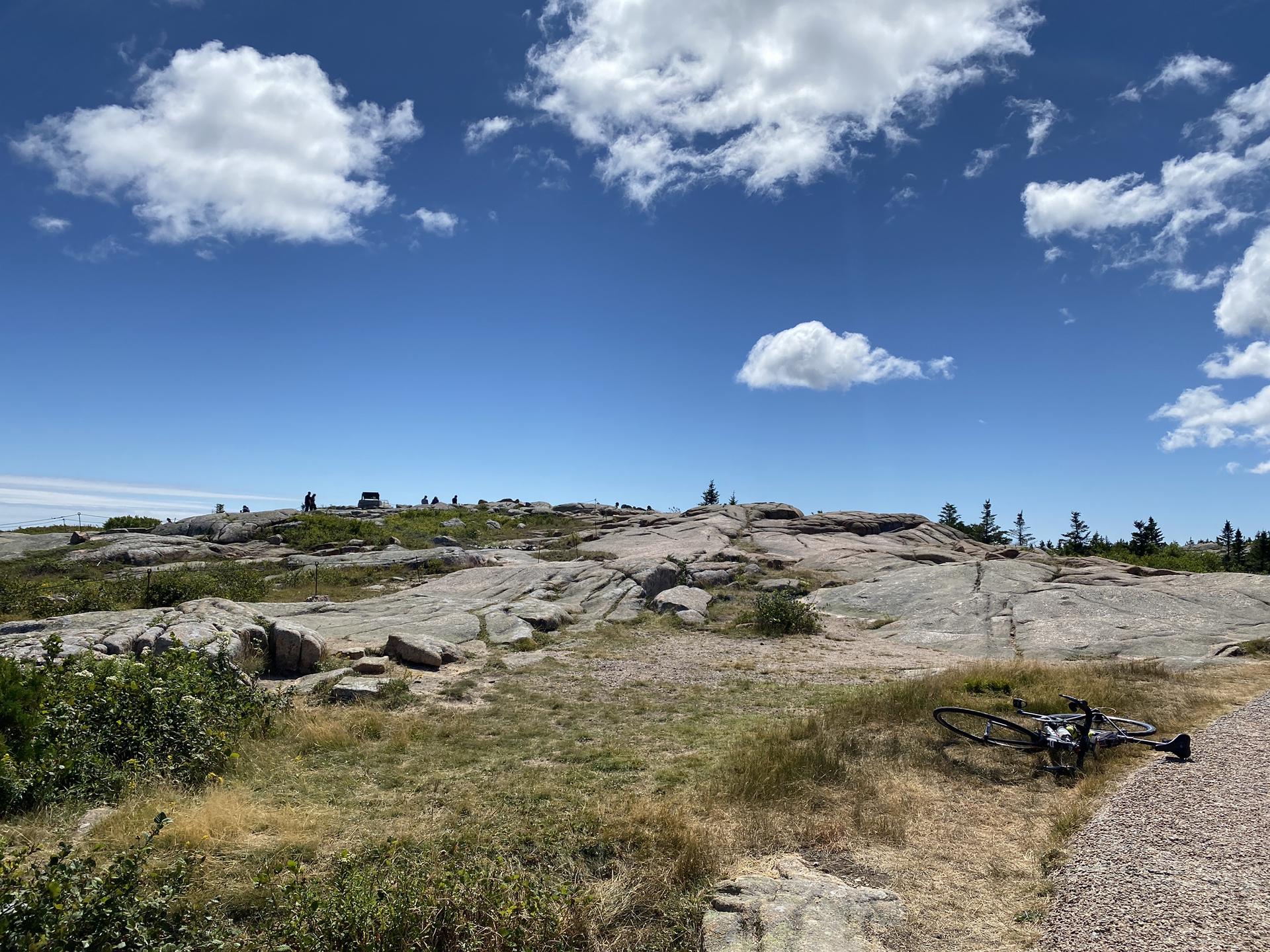 A bicycle rests on rocky, grassy terrain under a bright, cloudy sky.