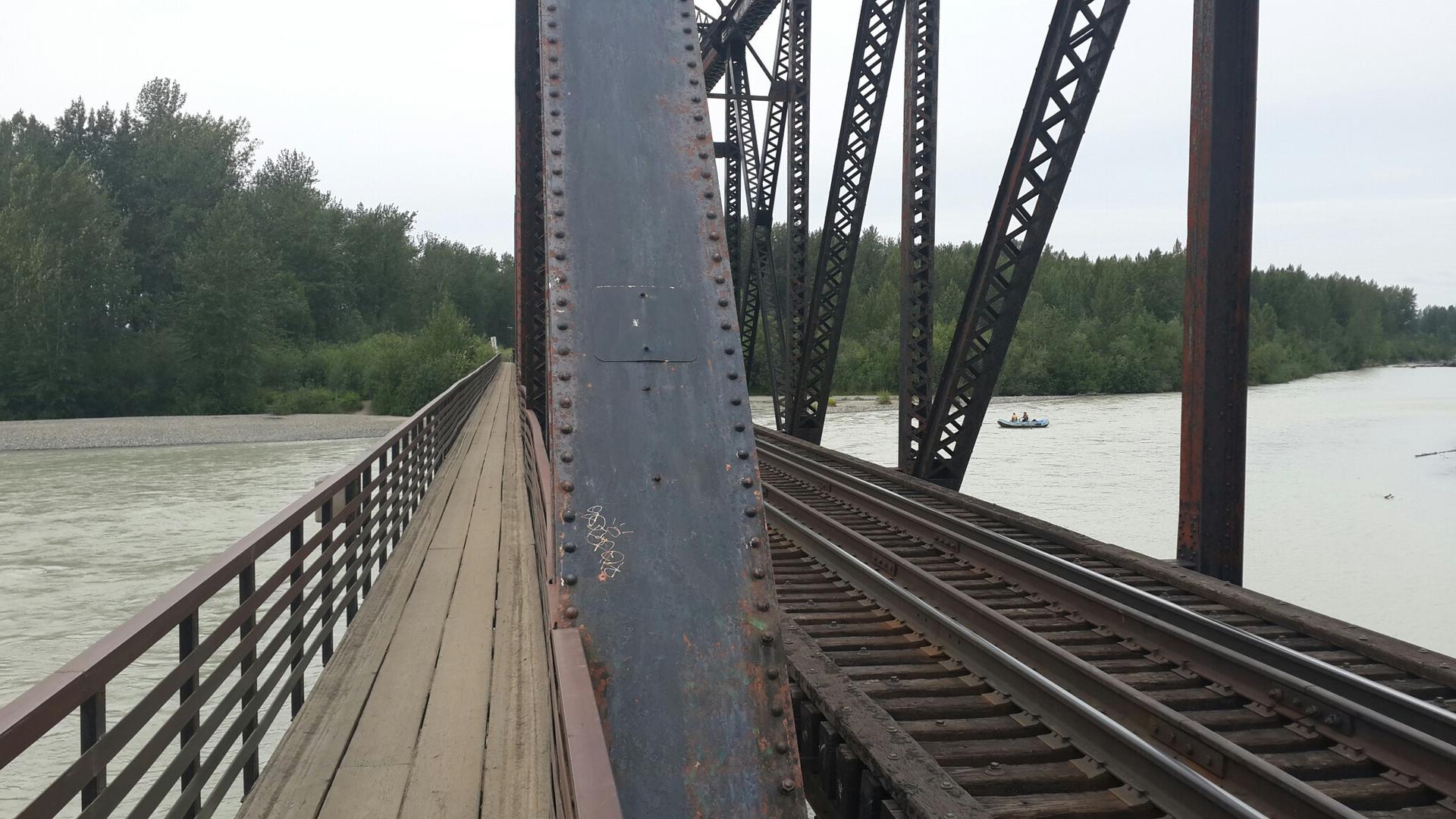 Close-up of a metal truss bridge with a wooden walkway over a murky river.