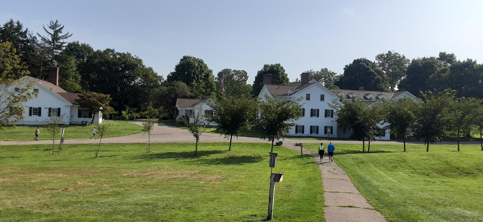 Two people walk on a paved path past white buildings and green fields.