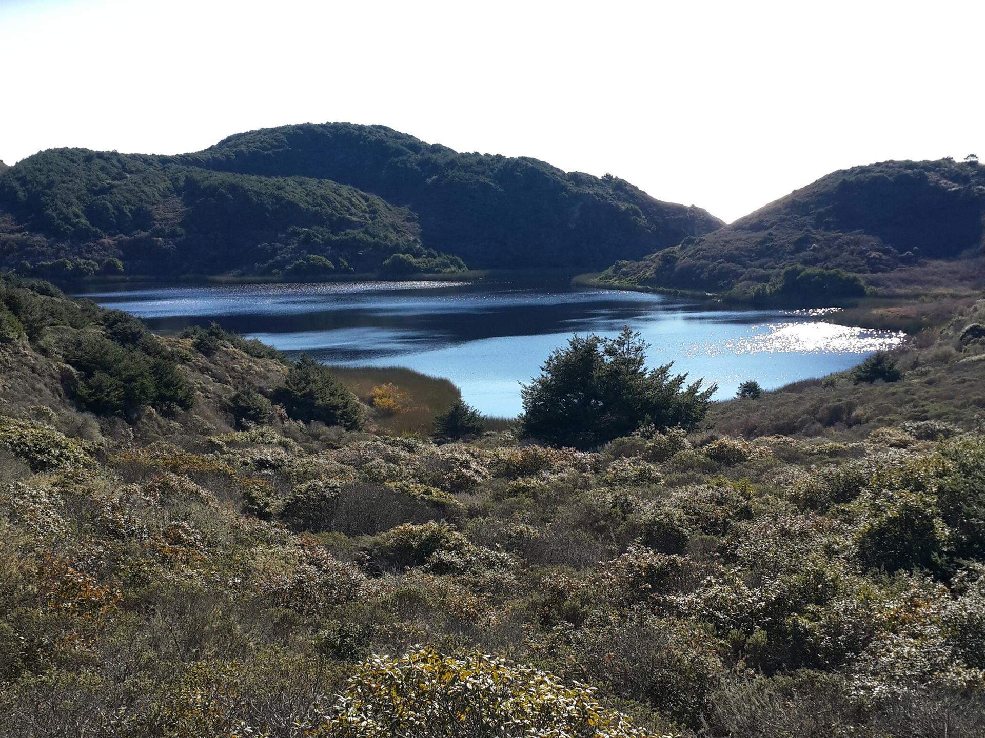 A shimmering lake surrounded by low, scrubby hills and dry vegetation in bright light.