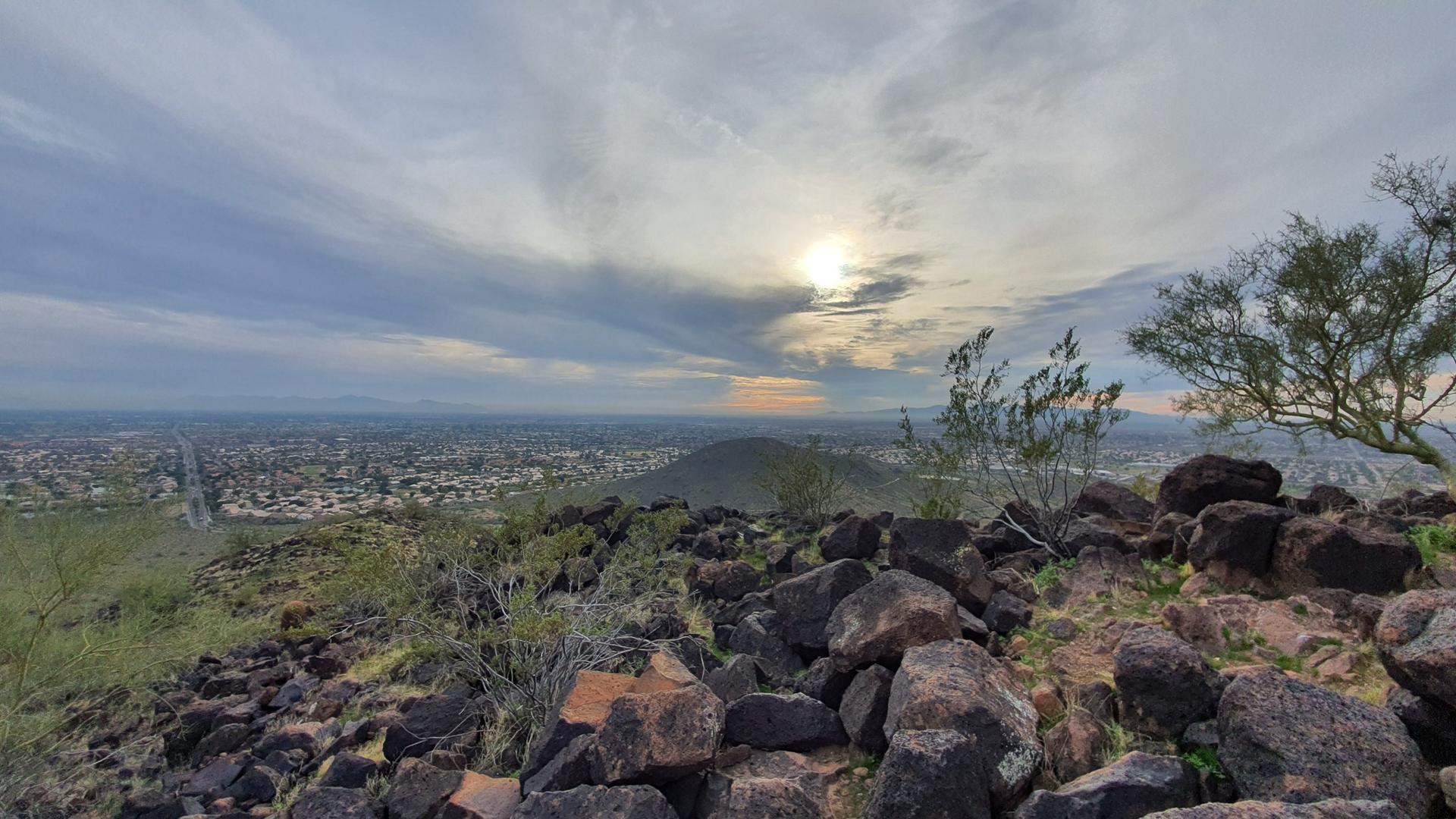 Rocky hilltop overlooking a city, with sun breaking through cloudy sky.