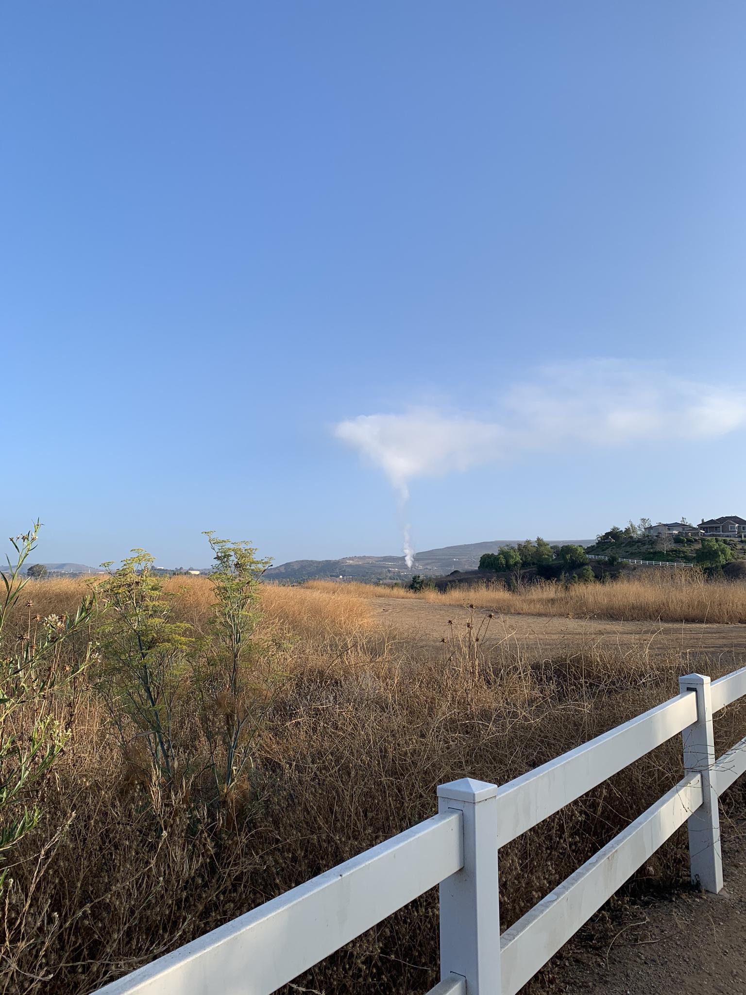 White fence, dry golden fields, and distant hills under a bright blue sky.