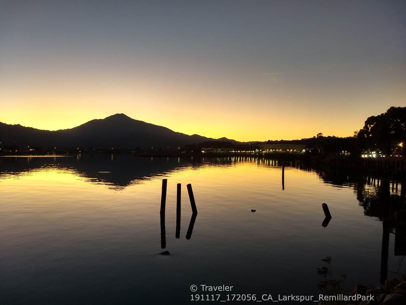 Golden sunset over a calm lake, reflecting silhouetted mountains and distant lights.