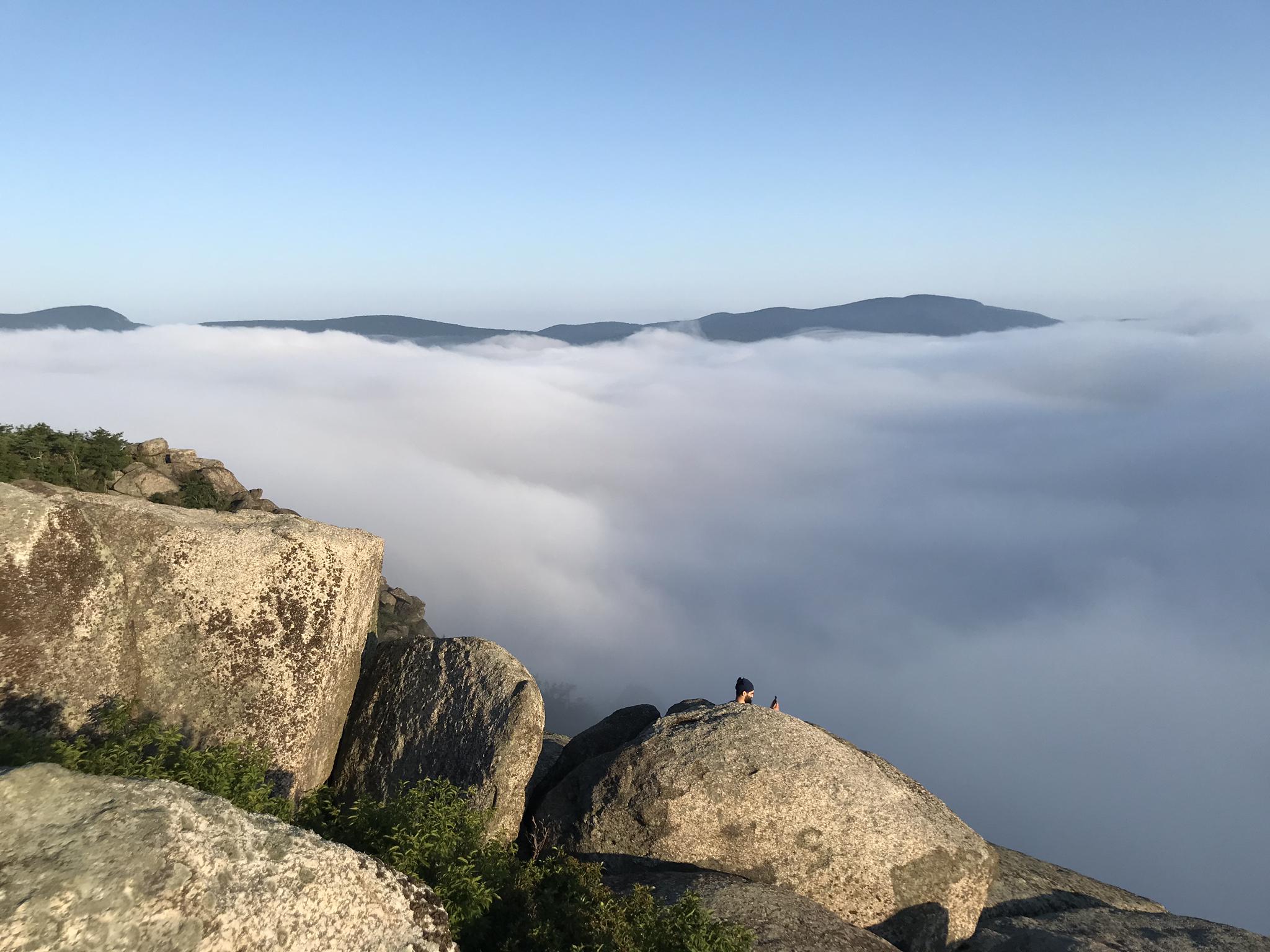 Person on rocky peak overlooking cloud inversion and distant mountains under clear sky.