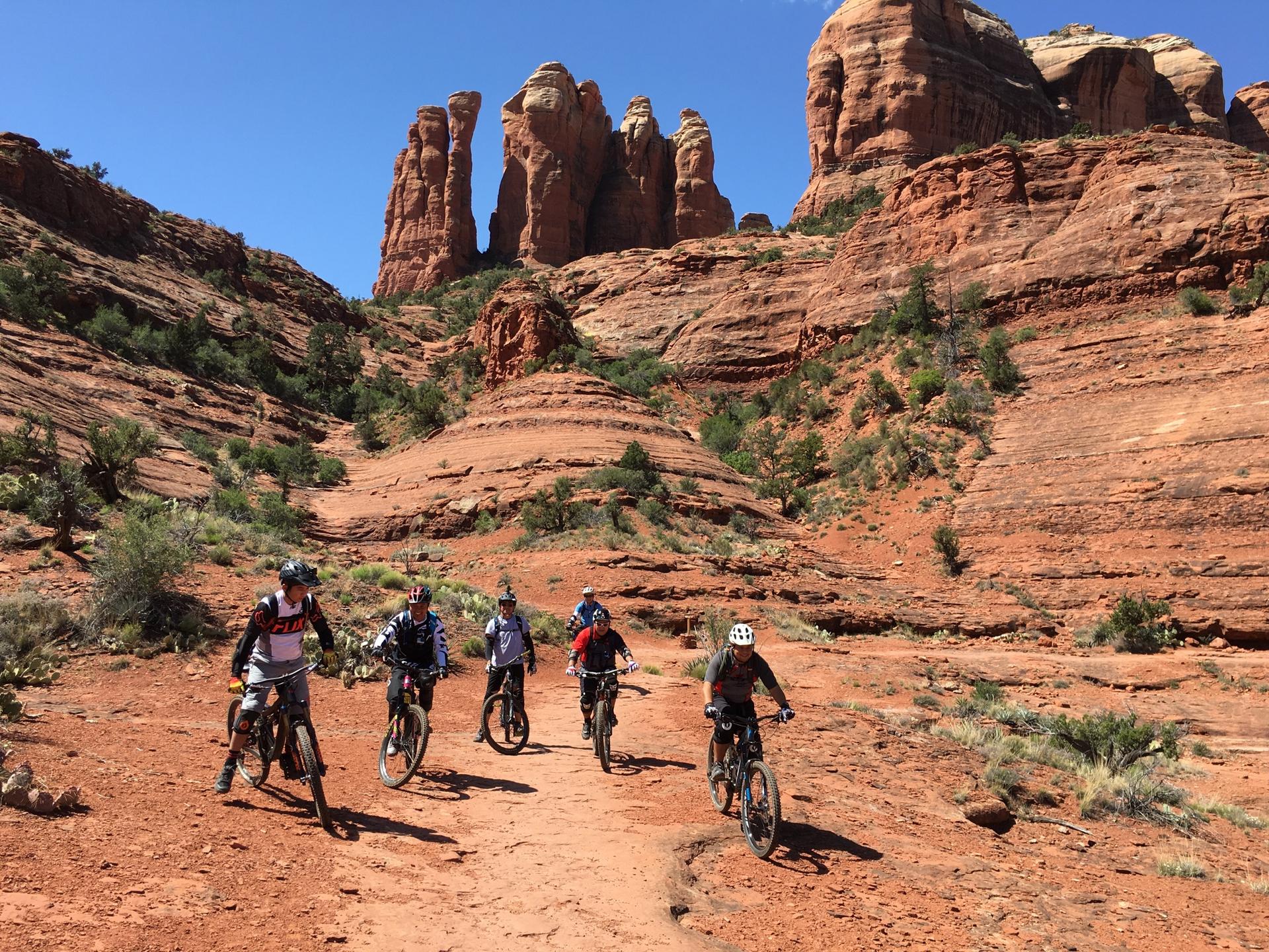 Five people mountain biking on a red dirt path among towering red rocks.