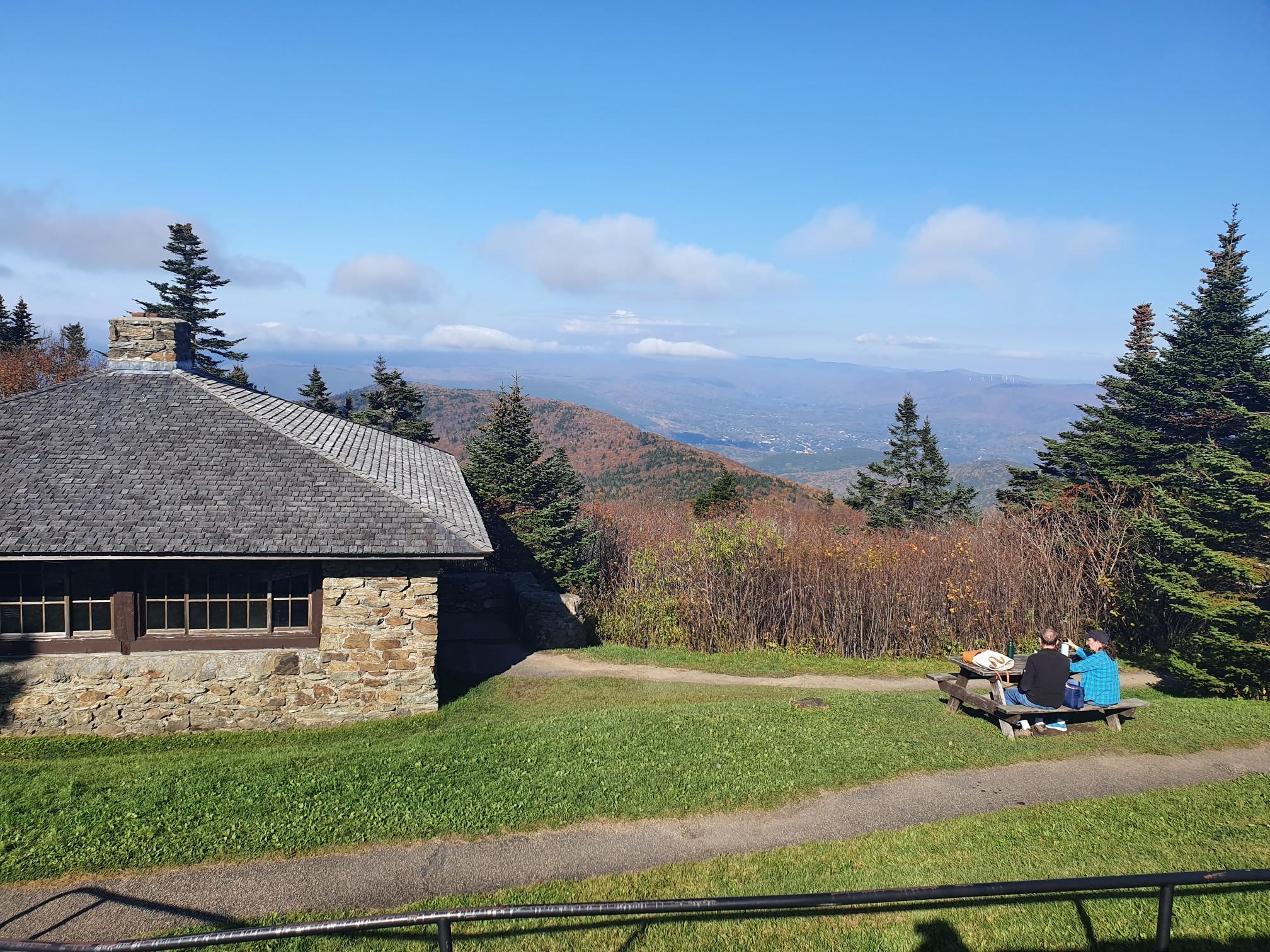 Stone building and people at a picnic table overlooking a mountain vista.