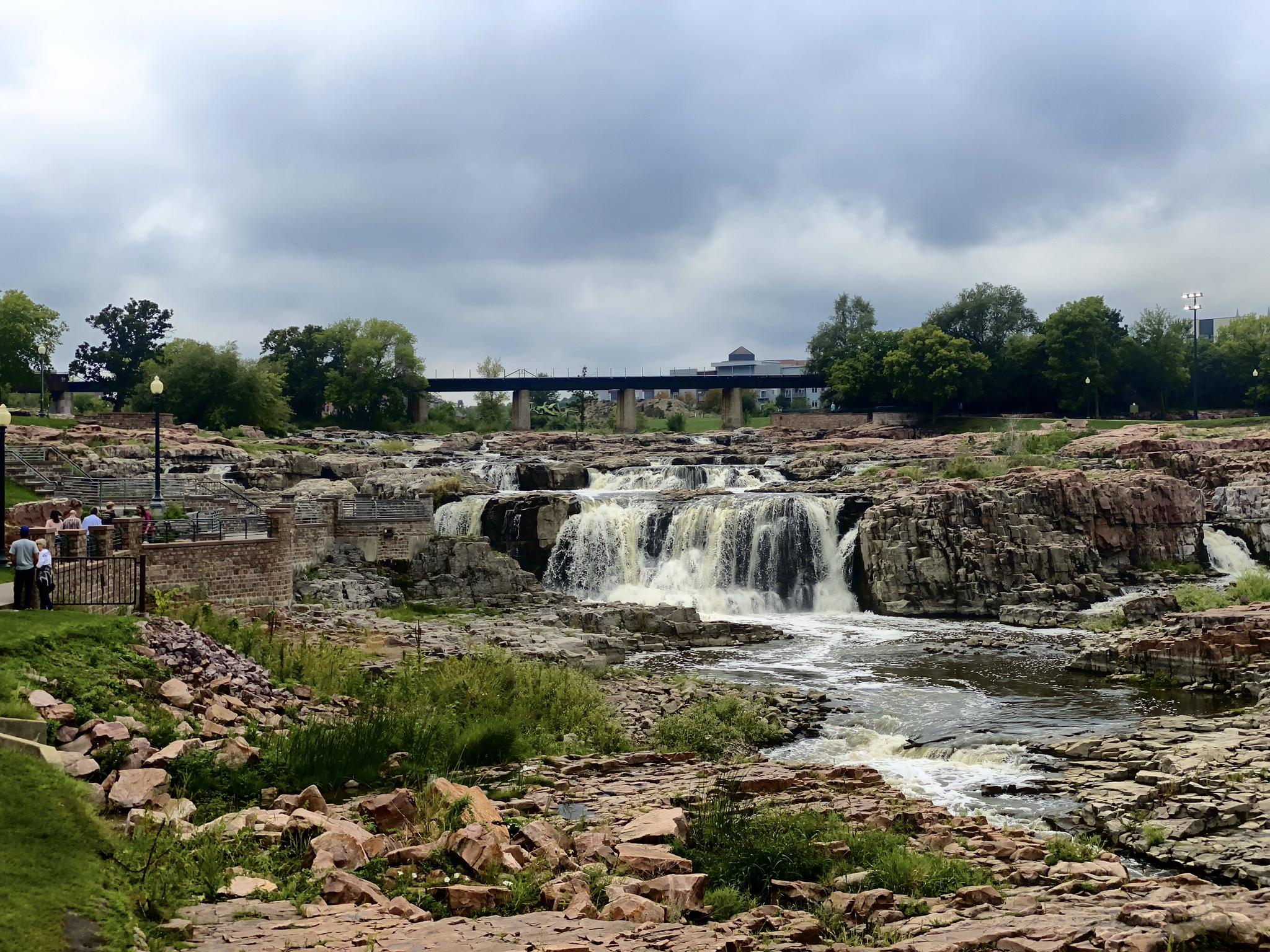 Powerful waterfall cascades over rocky riverbed under an overcast sky, people watching.