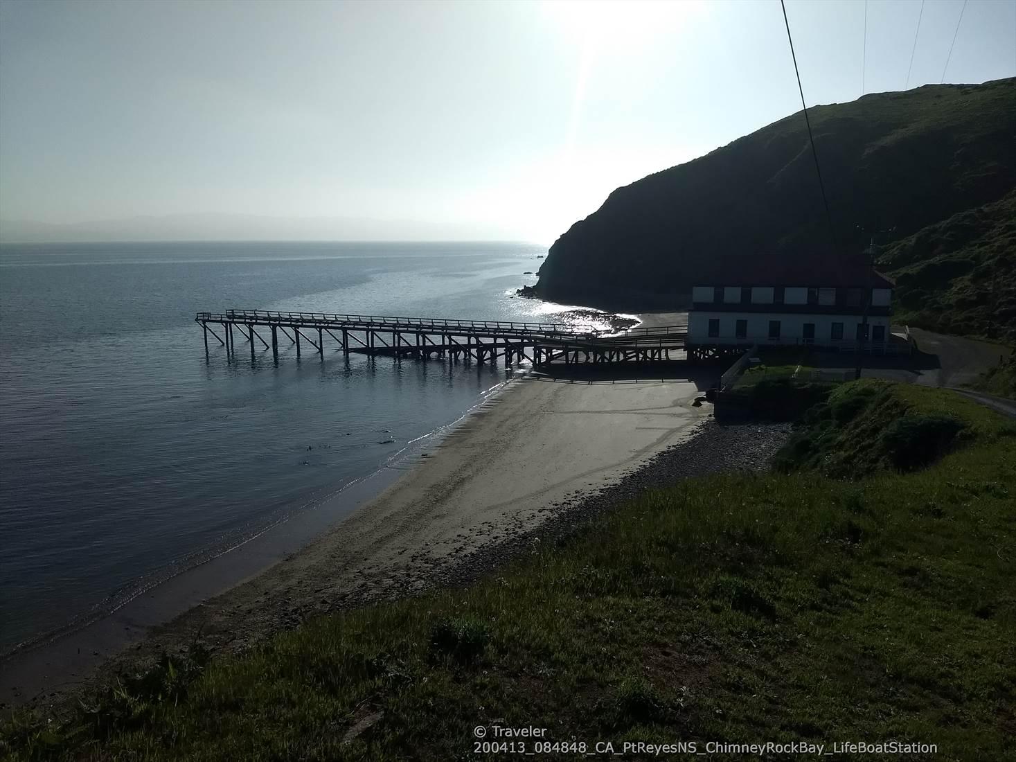A long pier extending into calm water next to a building on a beach, hazy sunlight.
